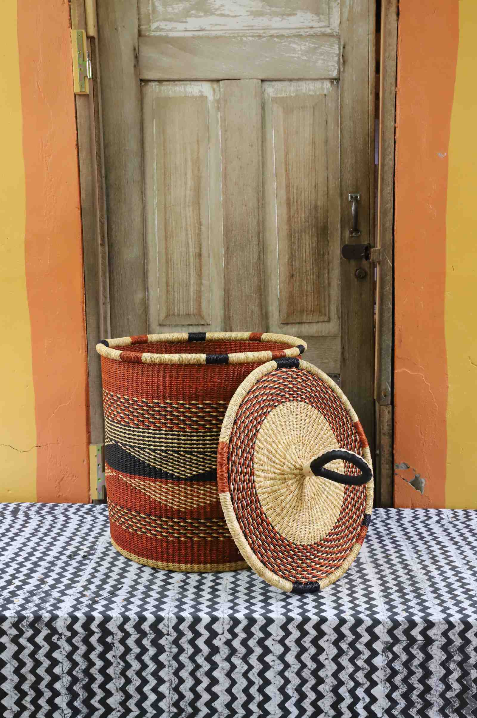 Handwoven Bolga laundry basket with lid removed, rust-orange body with dark wave motif and matching lid.