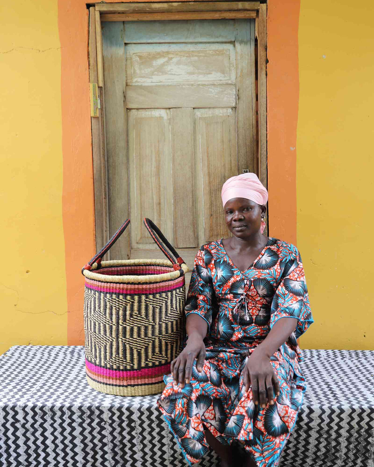 Small handwoven Bolga laundry basket in natural with red bands and dark weave, beside a seated woman.