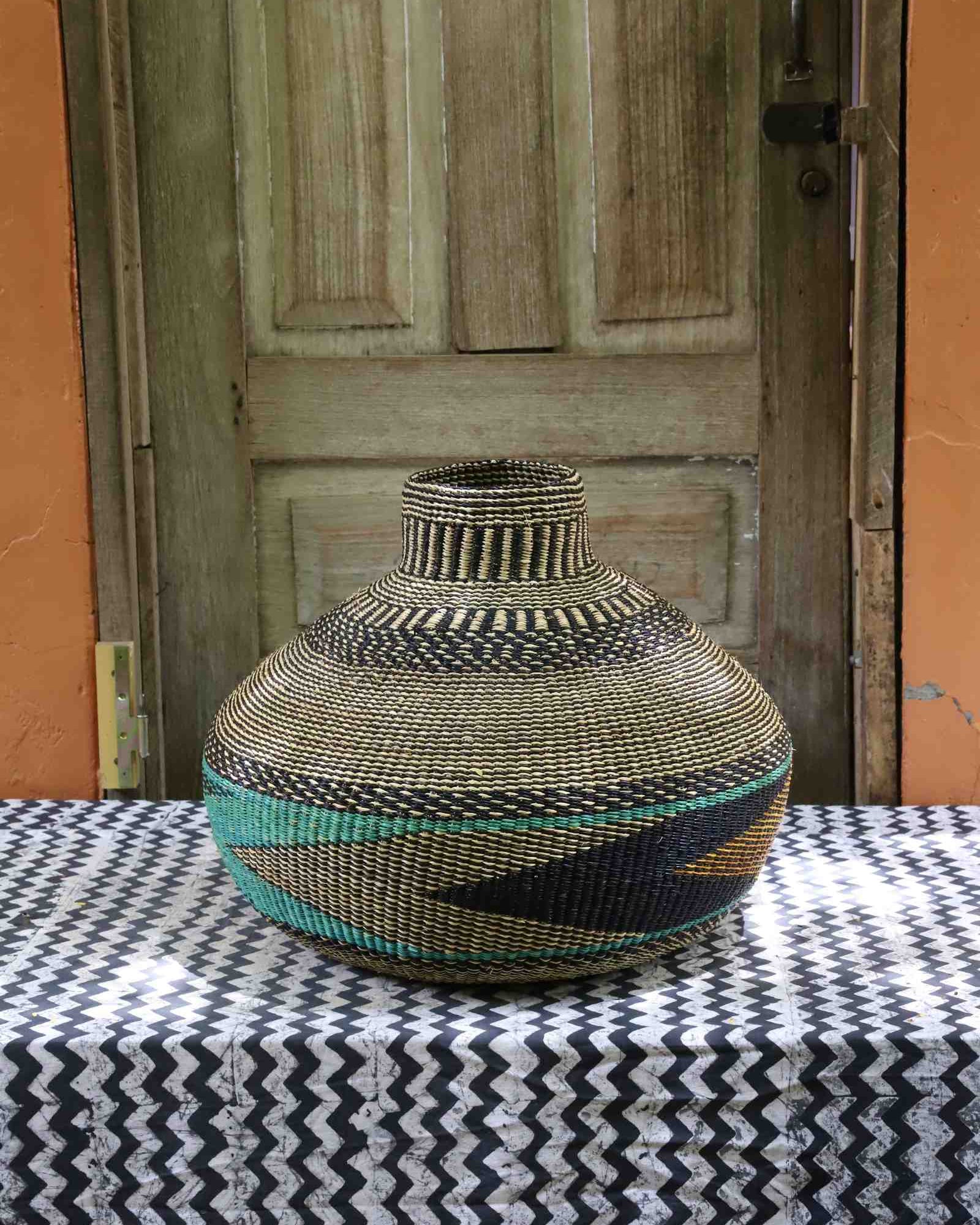 Handwoven African art basket in vase form, natural straw with black and teal geometric pattern, on a chevron surface.