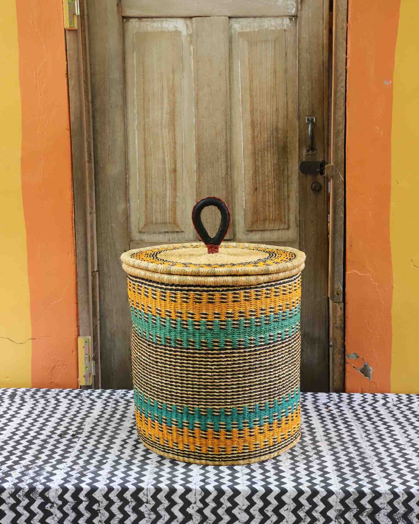 Handwoven Bolga laundry basket with coiled rim and lid in natural straw, striped in yellow-orange and teal.
