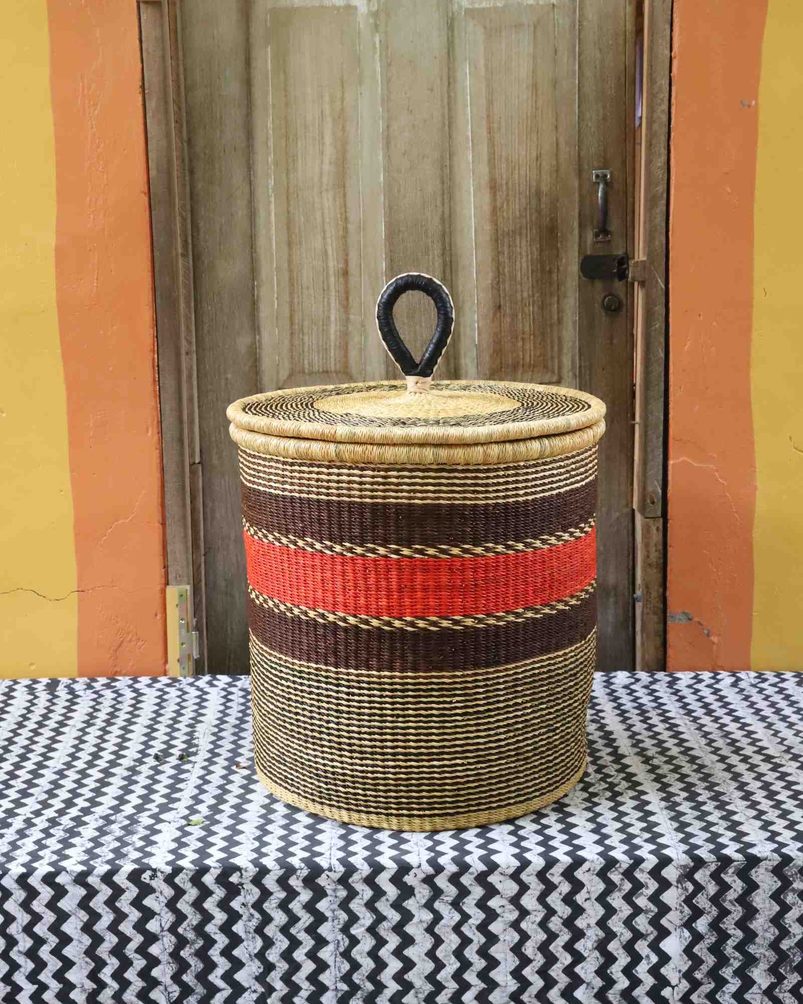 Large coiled-rim lidded laundry basket in natural straw with a red and brown band, styled on a chevron cloth.