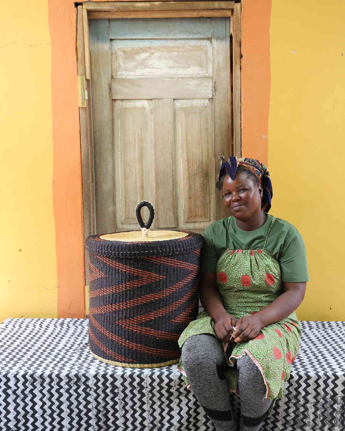 Handwoven Bolga laundry basket in dark blue with rust-orange zigzag, beside a seated woman in green.
