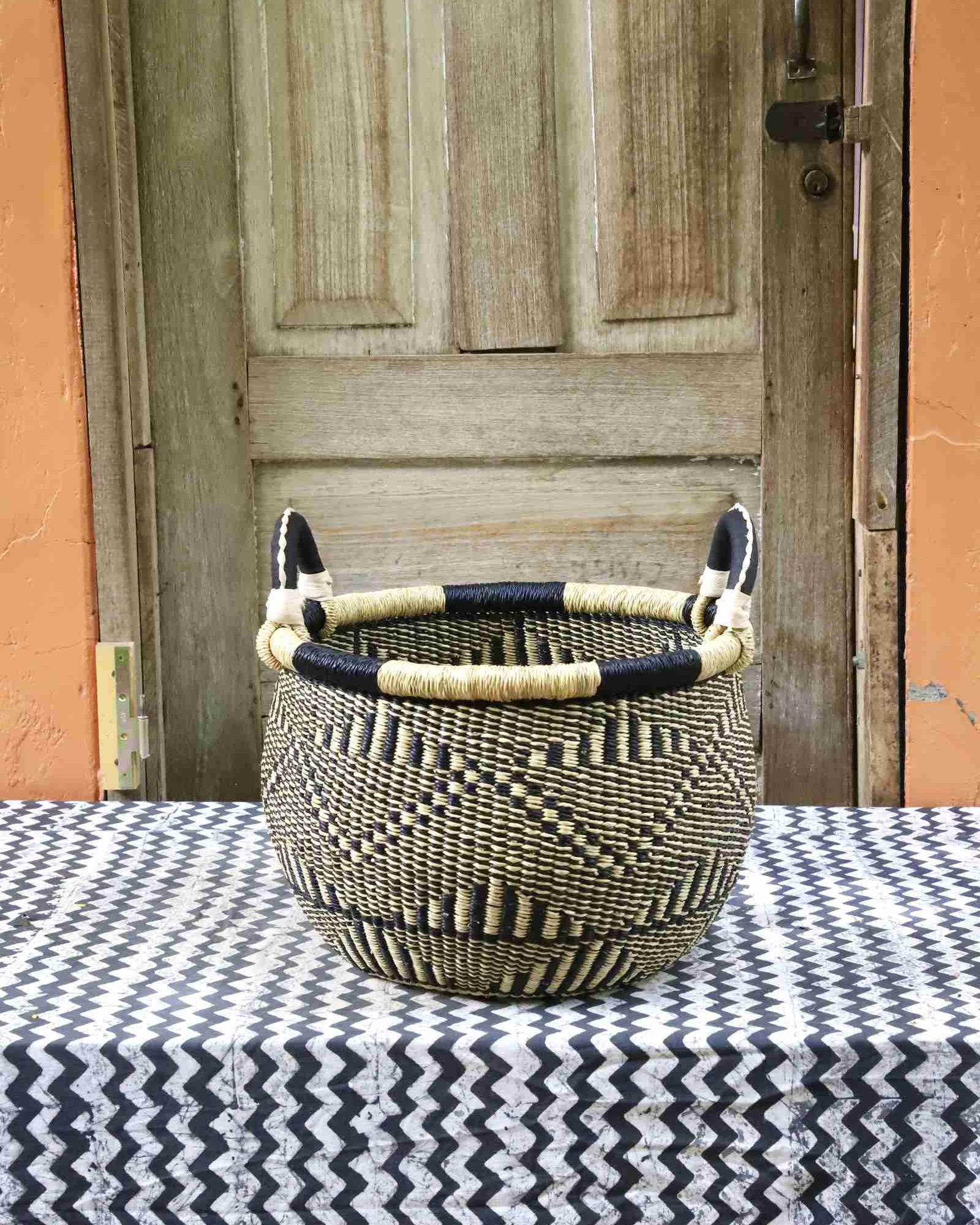 Handwoven Bolga storage basket in natural straw with black diamond pattern and leather loop handles, on a chevron surface.
