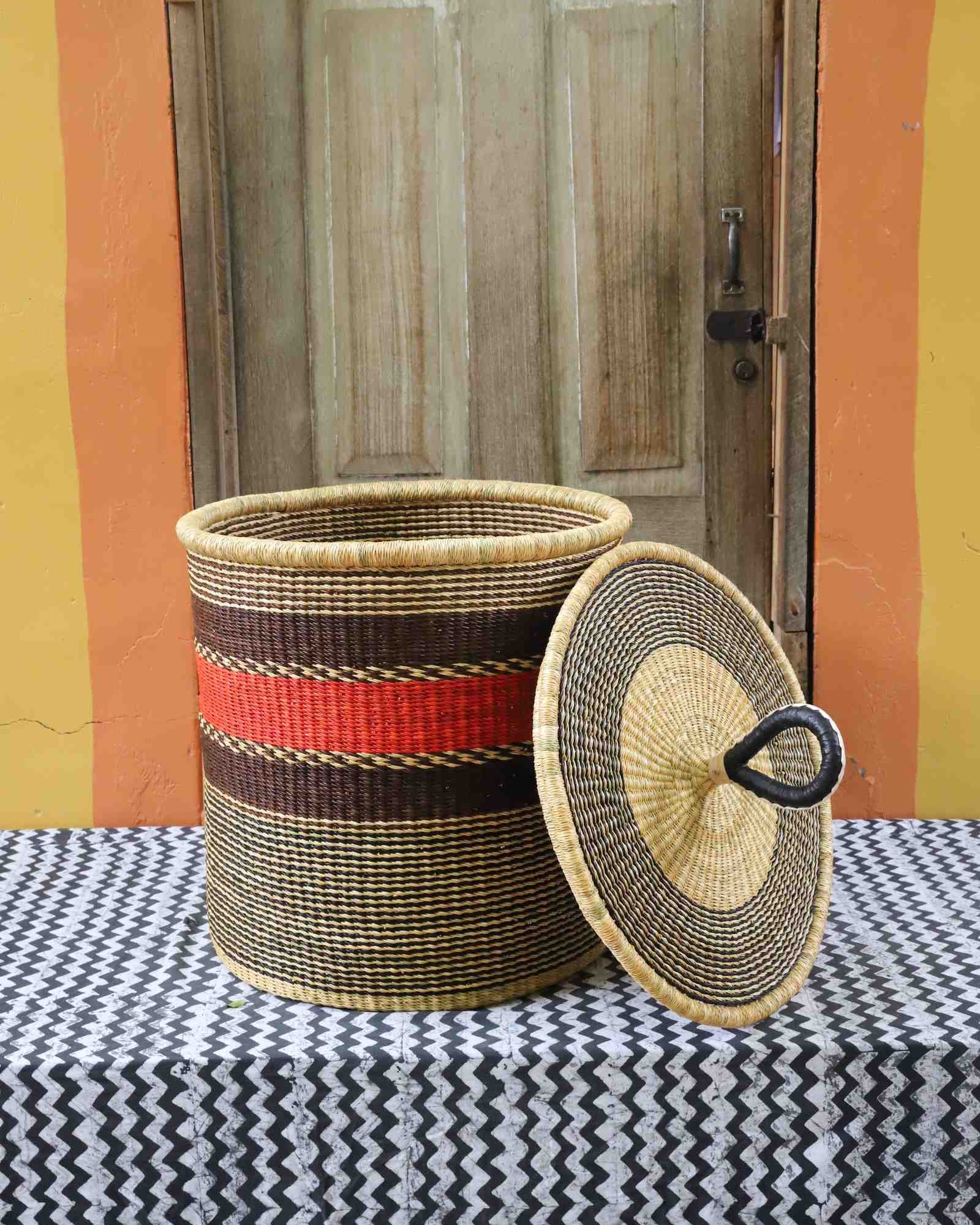 Large coiled-rim laundry basket in natural straw with a red and brown band, lid resting alongside on a chevron cloth.