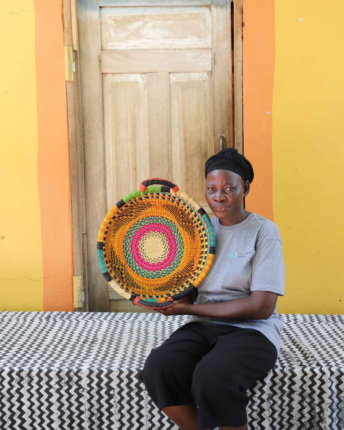 Handwoven Bolga wall tray disc with fine multicolour bands, held by a seated woman in grey.