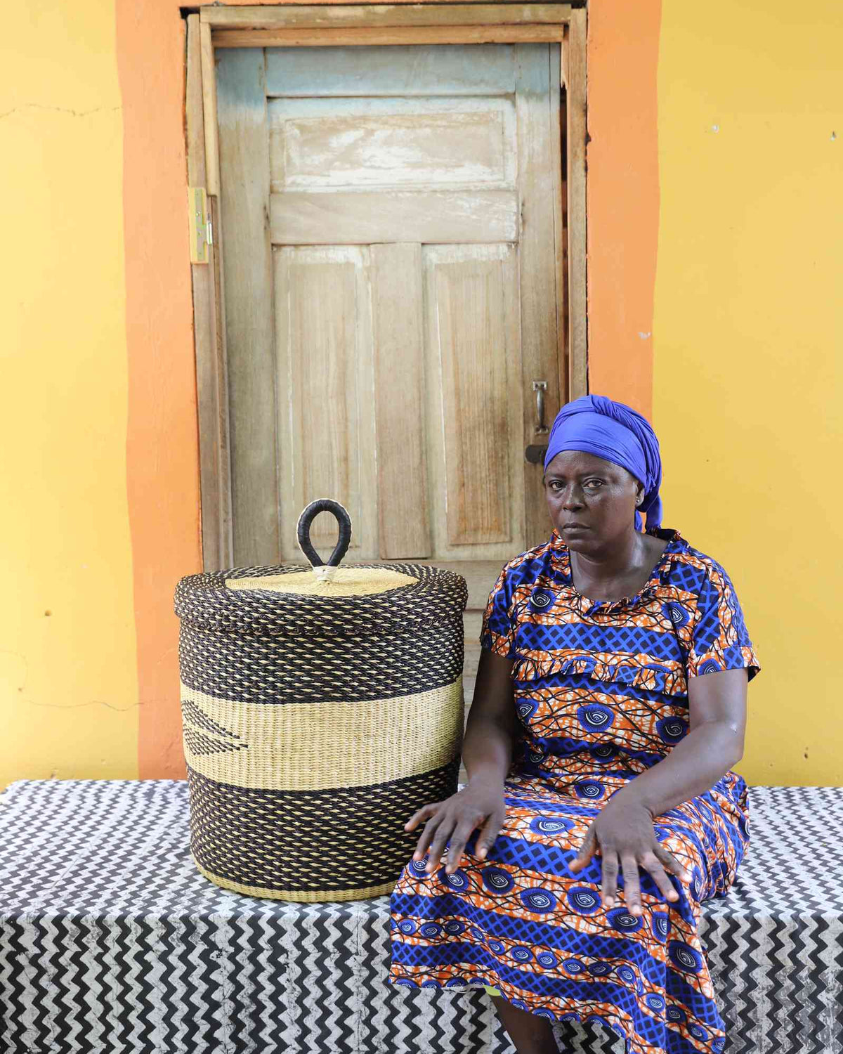 Handwoven Bolga laundry basket in dark straw with natural band, beside a seated woman in a printed dress.