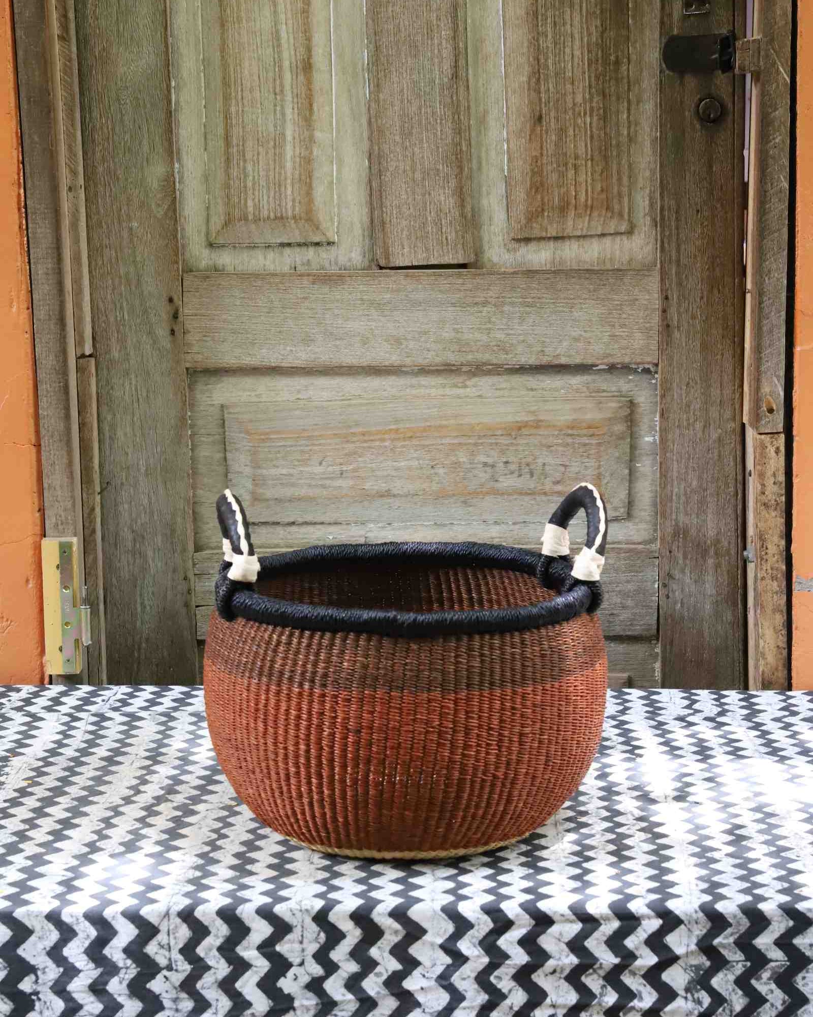 Handwoven Bolga storage basket in rust with black rim and leather loop handles, styled on a chevron surface.
