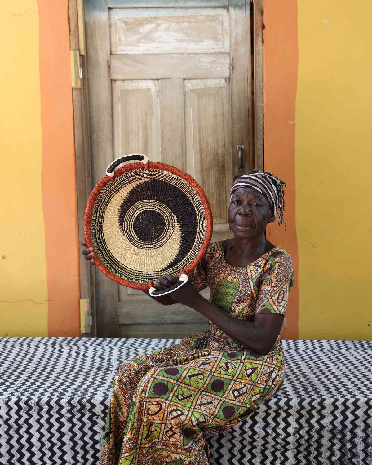 Handwoven Bolga wall tray disc with orange rim and black rings, held by a seated woman in green.