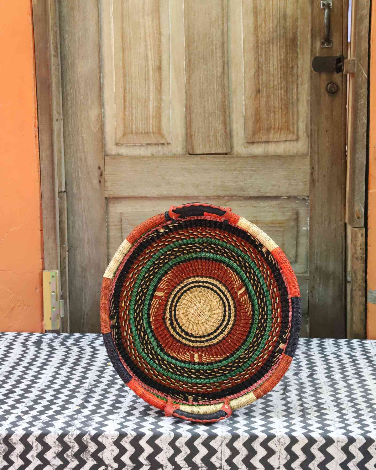 Handwoven Bolga wall tray disc with concentric orange, black, red and green banded rings.