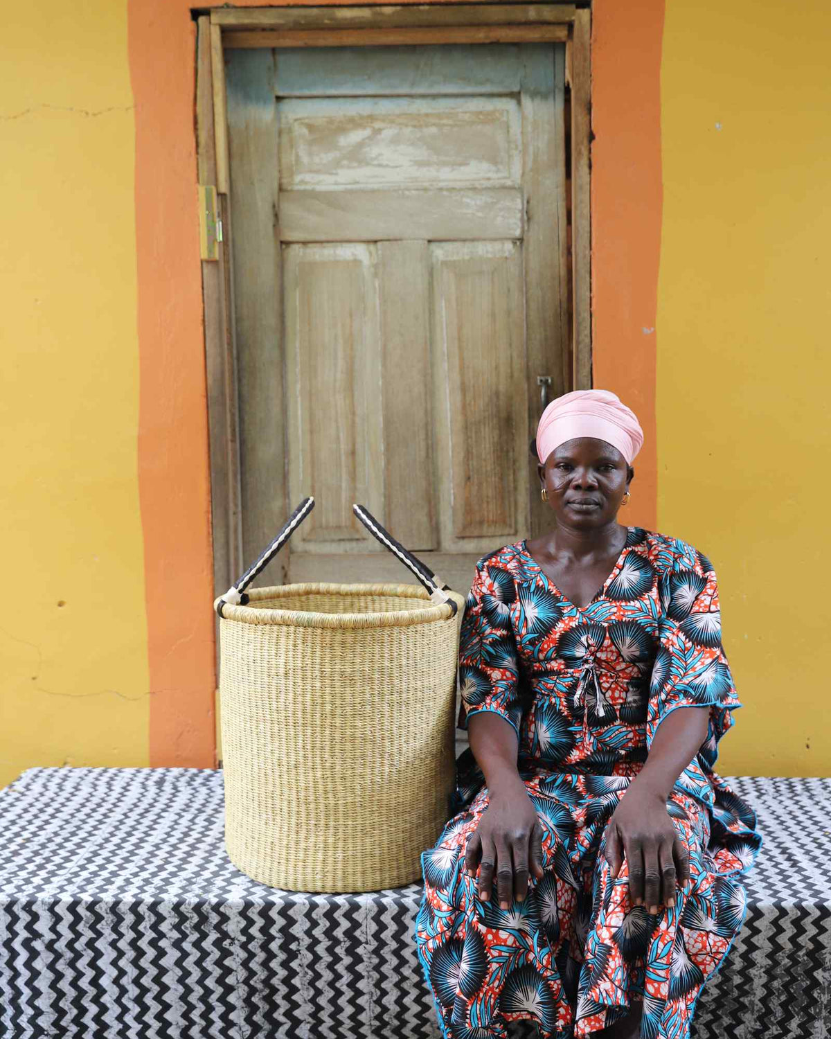 Small handwoven Bolga laundry basket in plain natural straw, beside a seated woman in a printed dress.