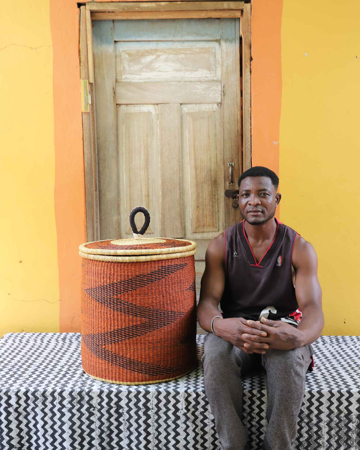 Handwoven Bolga laundry basket in dyed rust-orange with dark zigzag, beside a seated man in a dark vest.