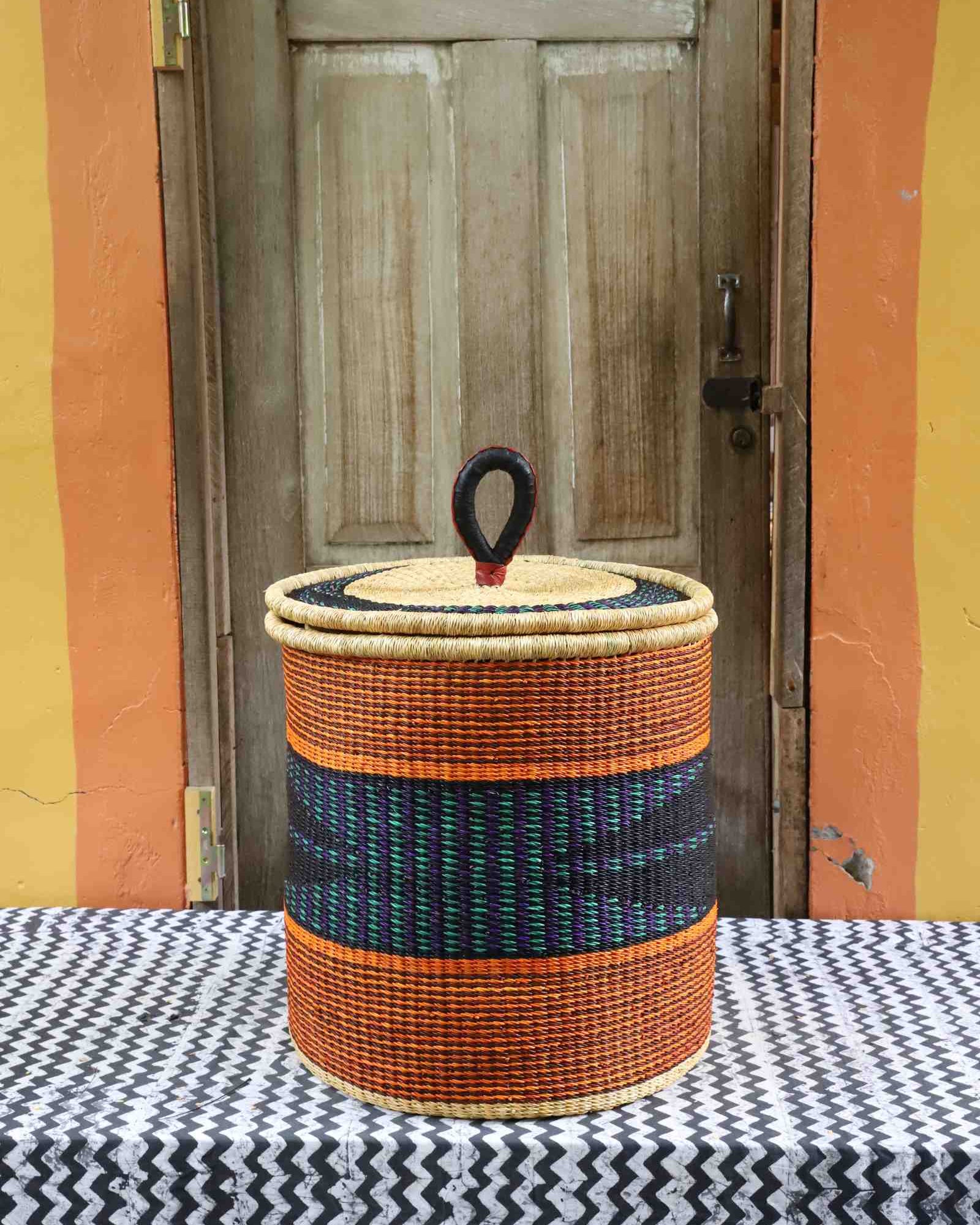 Handwoven Bolga laundry basket with coiled rim and lid in natural straw with orange bands and a dark blue centre band.