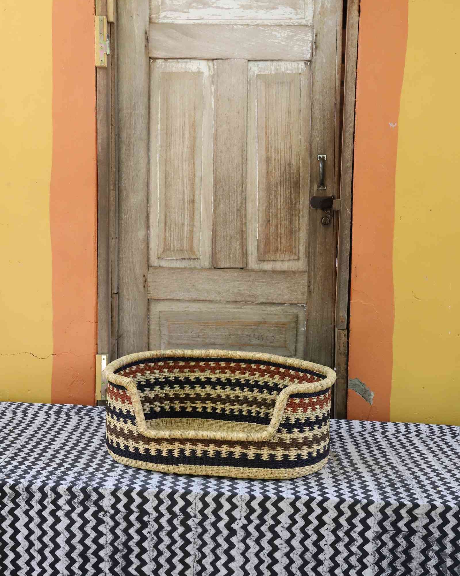 Small handwoven Bolga pet bed in natural straw with a black and orange zigzag pattern around the rim.