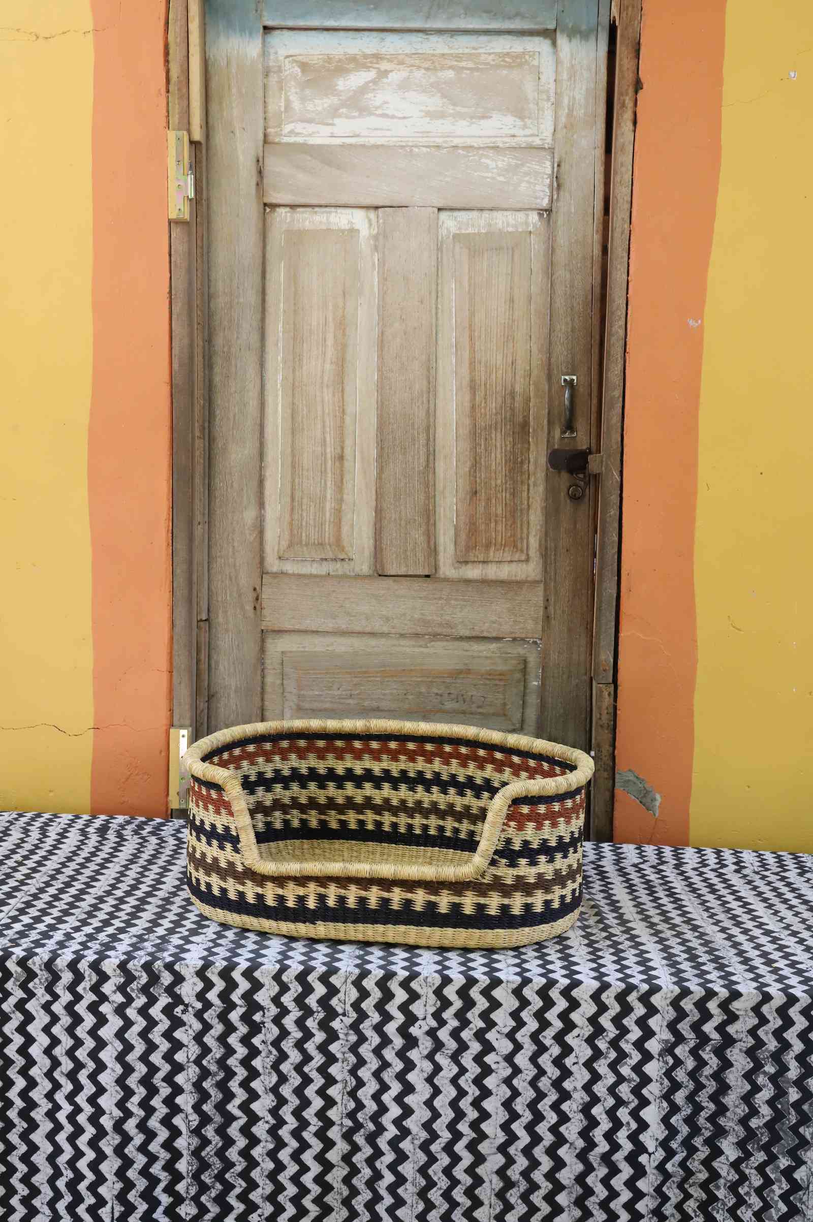 Small handwoven Bolga pet bed in natural straw with a black and orange zigzag pattern around the rim.