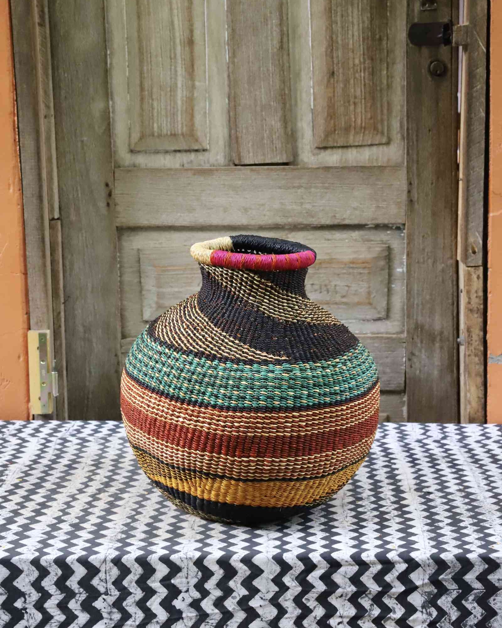 Handwoven African décor basket with horizontal bands of gold, terracotta, teal and black, on a chevron surface.