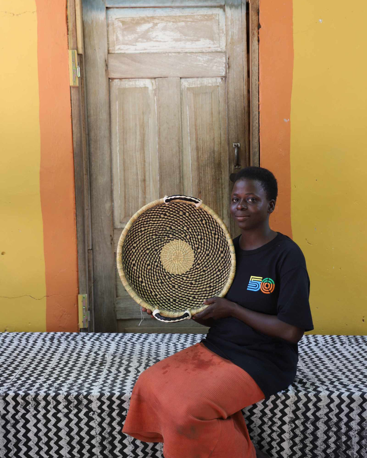 Handwoven Bolga wall tray disc in dark straw with yellow centre, held by a seated woman in dark top.