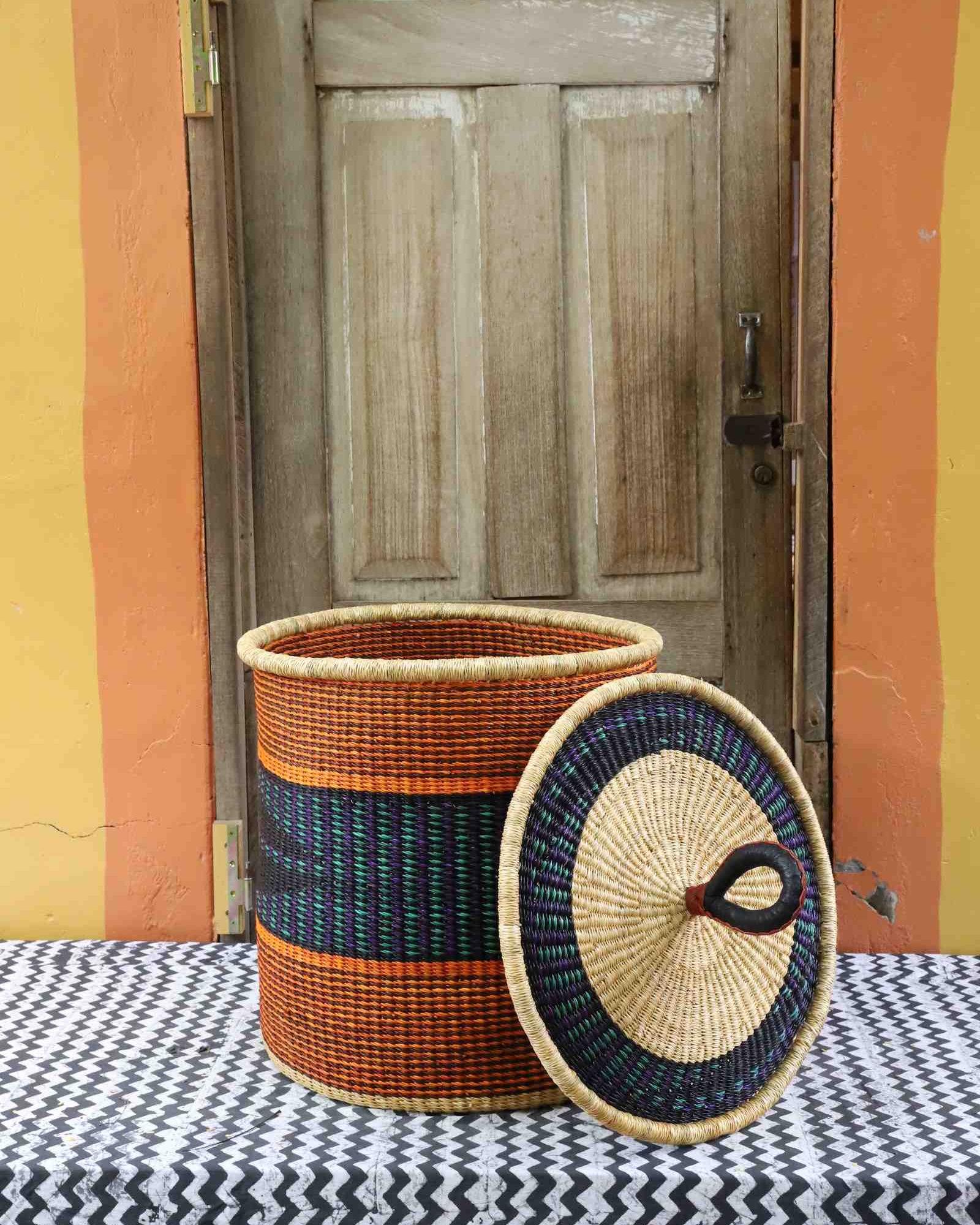 Handwoven Bolga laundry basket with lid removed, natural body with orange and dark blue bands and matching lid.