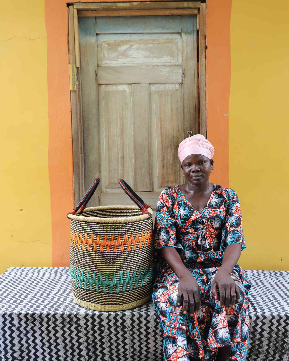 Small handwoven Bolga laundry basket in natural with orange and teal bands, beside a seated woman in floral.