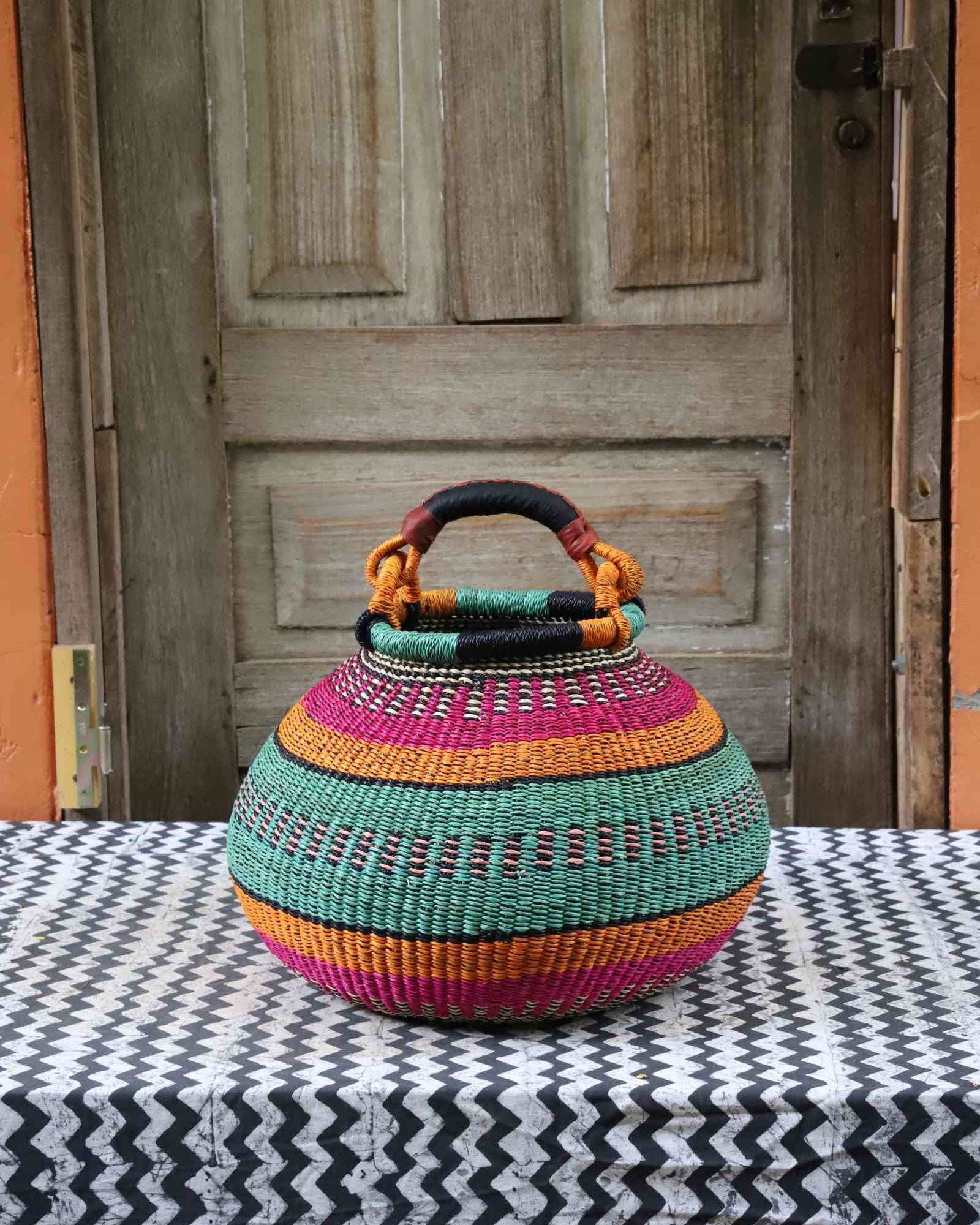 Large handwoven Bolga pot basket in bold bands of pink, teal and amber with a leather top handle, on a chevron surface.