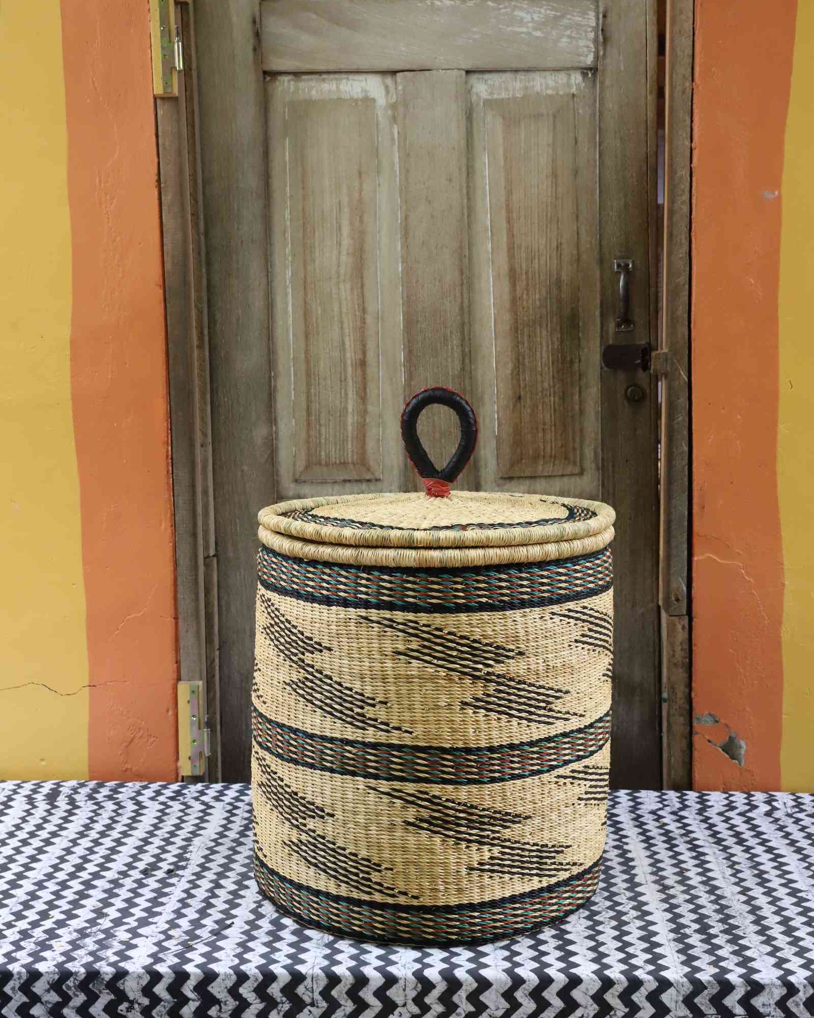 Handwoven Bolga laundry basket with coiled rim and lid in natural straw with fine black bands and wave motifs.
