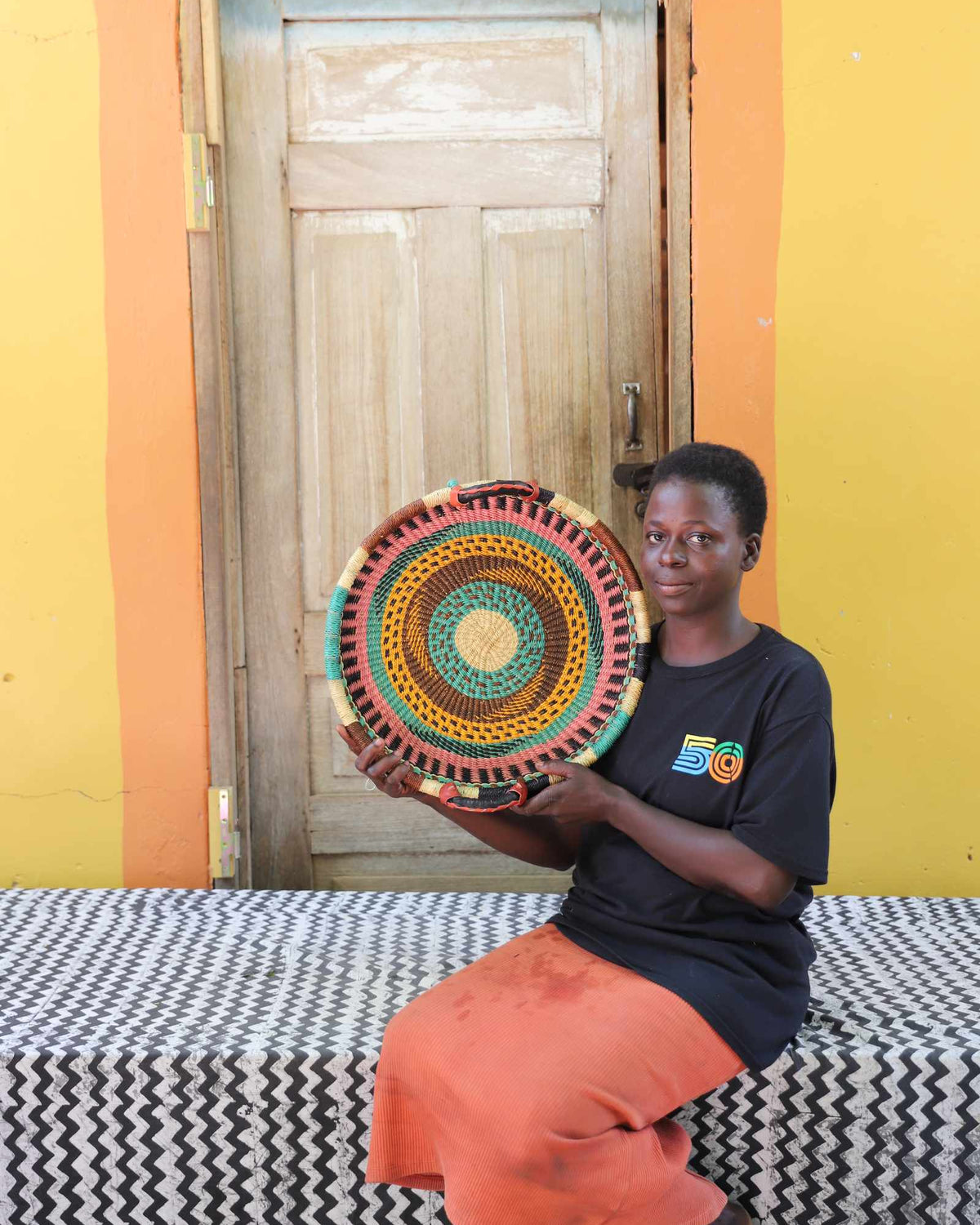 Handwoven Bolga wall tray disc with red and green bands, held by a seated woman in an orange skirt.