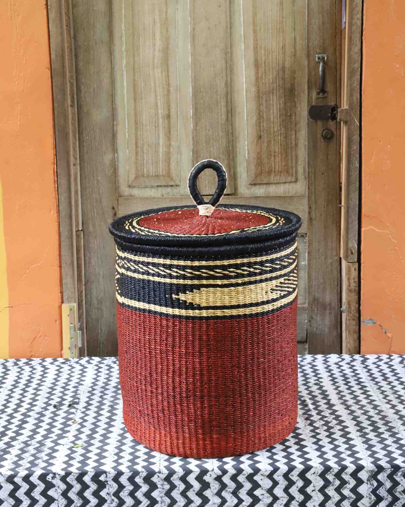Coiled-rim lidded laundry basket in solid rust weave with a black and tan banded rim, styled on a chevron cloth.