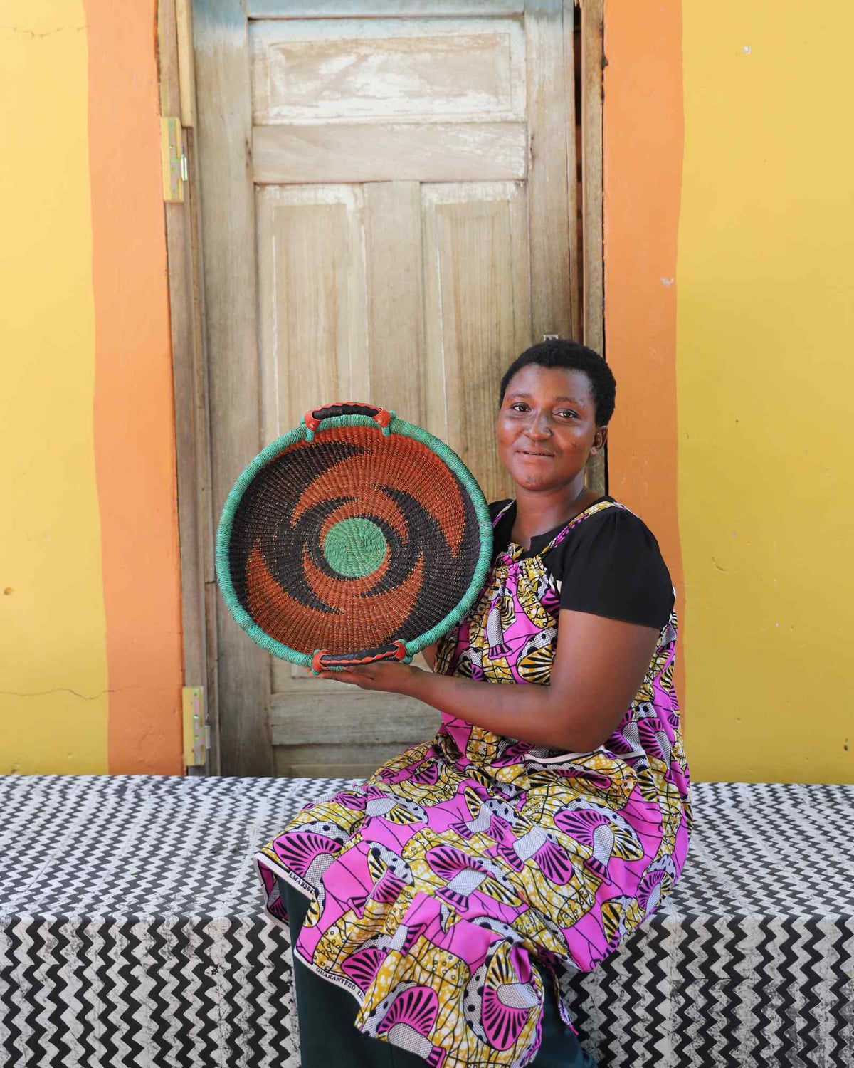 Handwoven Bolga wall tray disc with green rim and orange bands, held by a seated woman in floral.