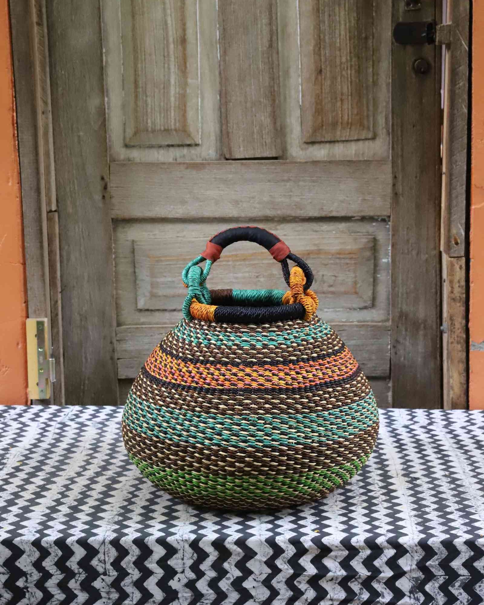 Handwoven Bolga pot basket in multicoloured bands of green, teal and amber with a leather top handle, on a chevron surface.