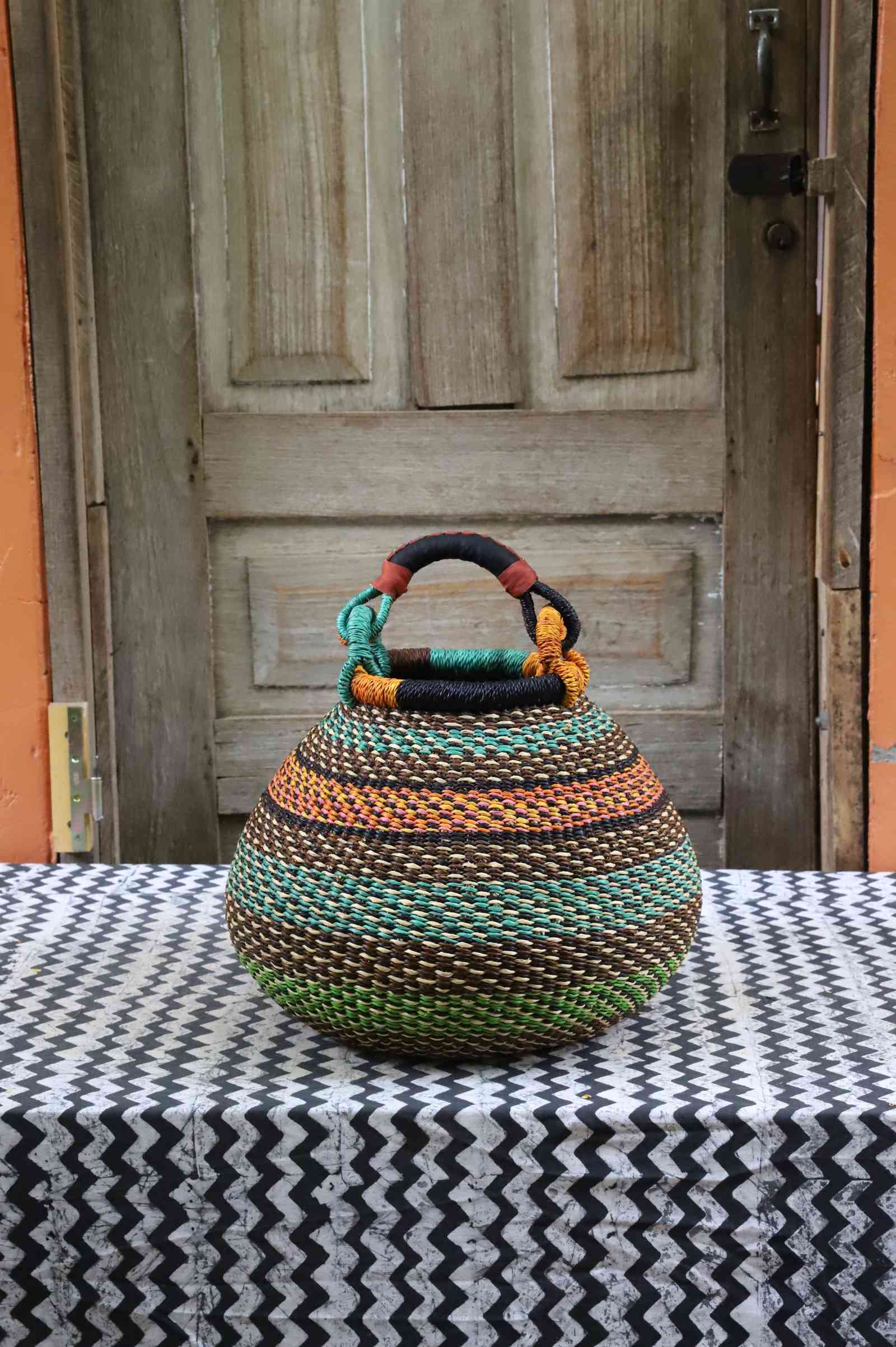 Handwoven Bolga pot basket in multicoloured bands of green, teal and amber with a leather top handle, on a chevron surface.