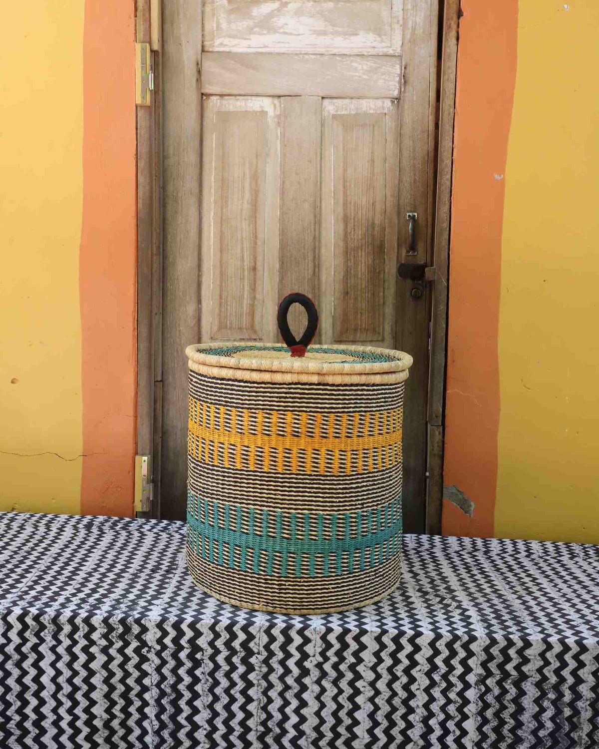 Handwoven Bolga laundry basket with coiled rim and lid in natural straw with yellow and teal banded stripes.