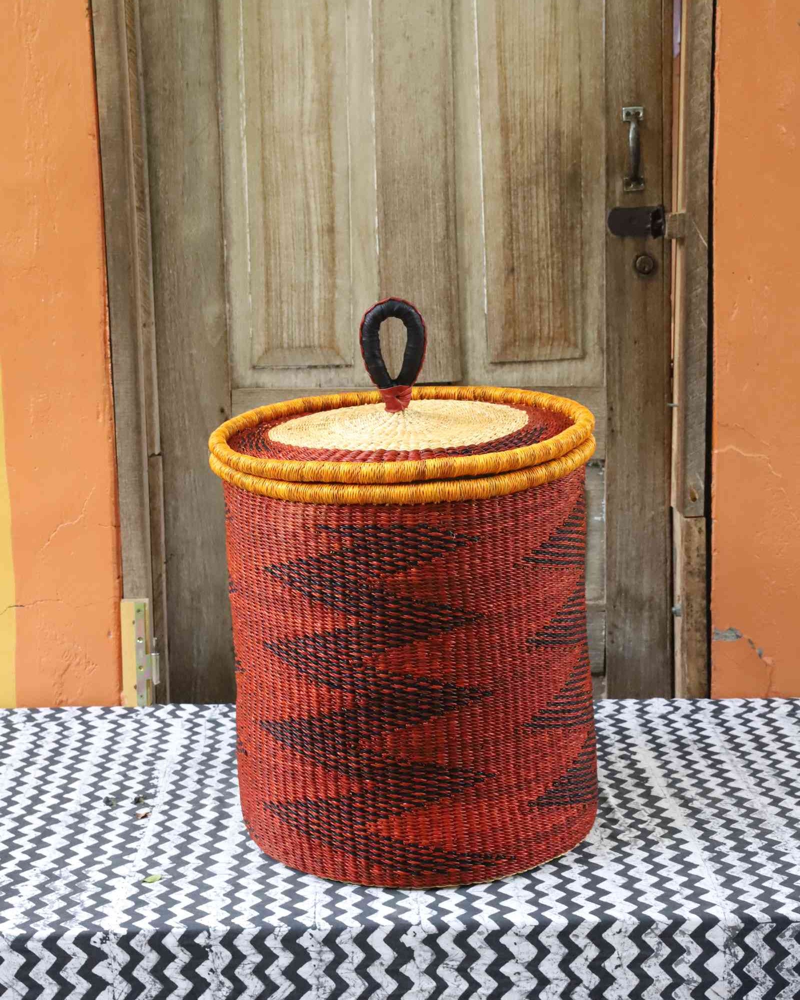 Coiled-rim lidded laundry basket in rust weave with a black diamond pattern and mustard rim, styled on a chevron cloth.