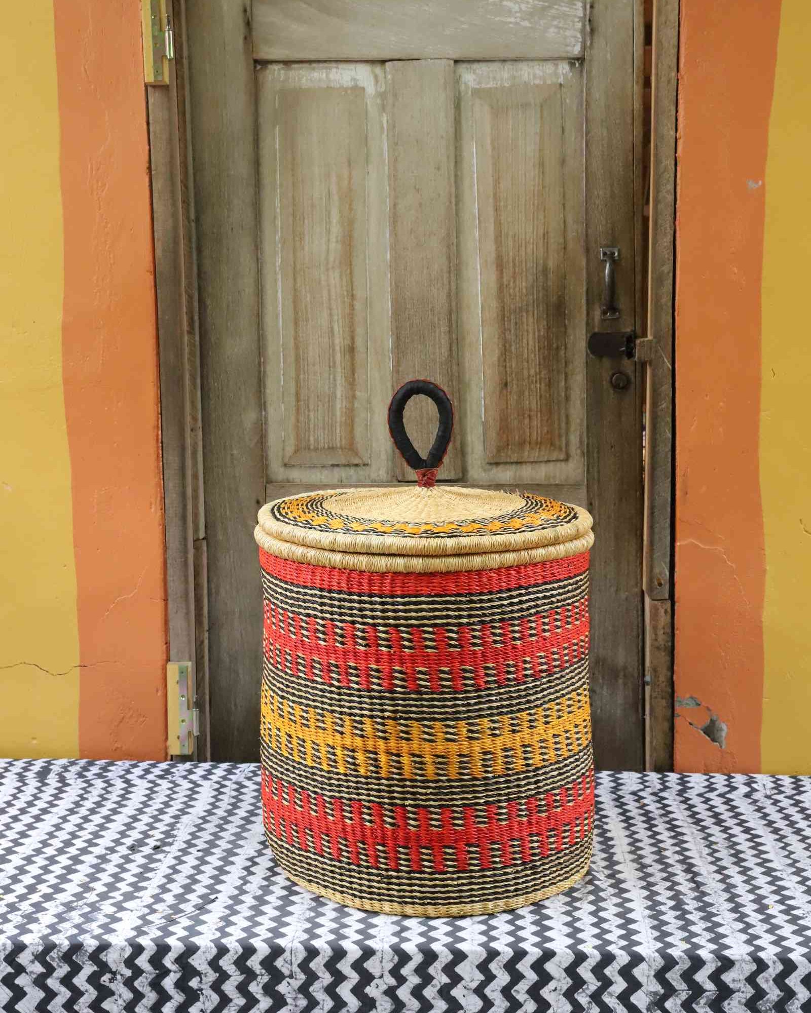 Handwoven Bolga laundry basket with coiled rim and lid in natural straw with red, orange and yellow banded stripes.