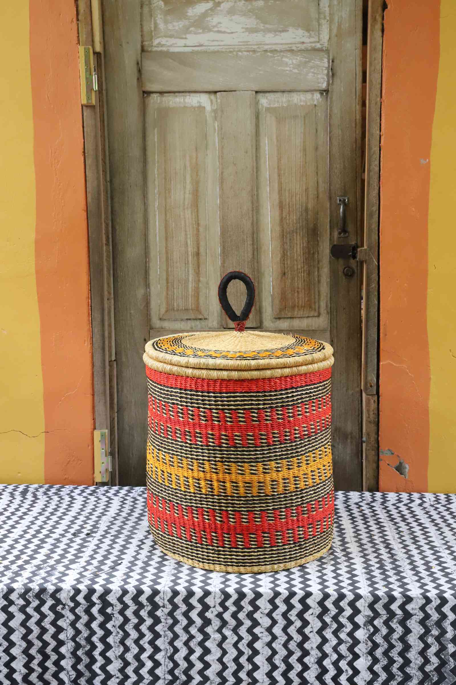 Handwoven Bolga laundry basket with coiled rim and lid in natural straw with red, orange and yellow banded stripes.