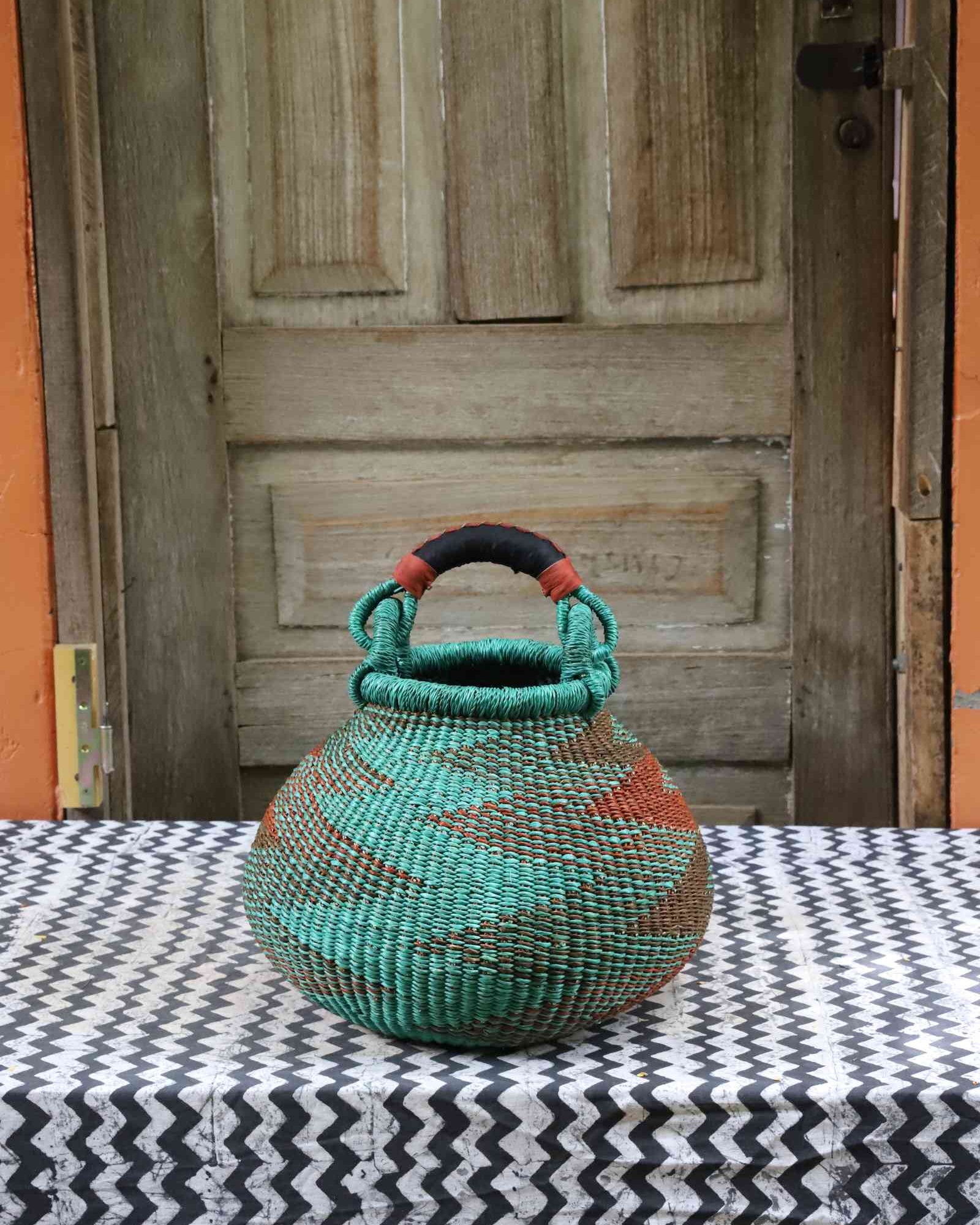 Small handwoven Bolga pot basket in teal and terracotta elephant grass with a leather top handle, on a chevron surface.