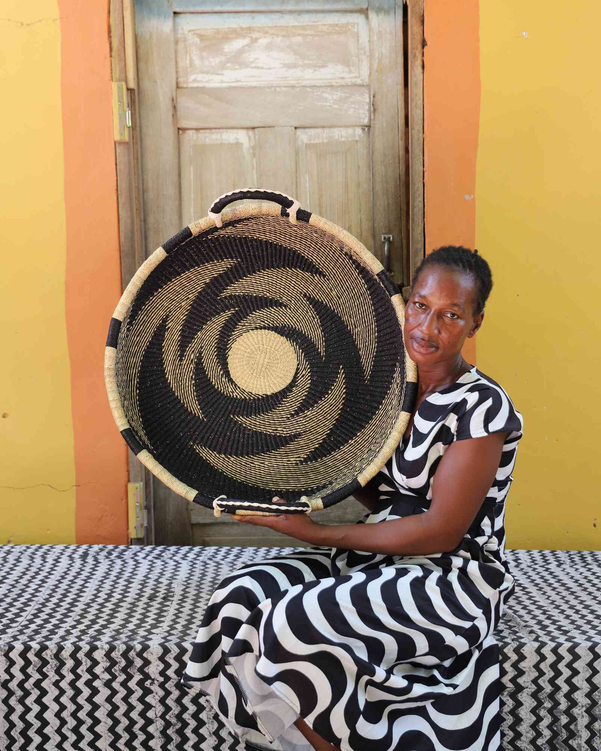 Medium handwoven Bolga wall tray disc with bold black pinwheel, held by a seated woman in stripes.