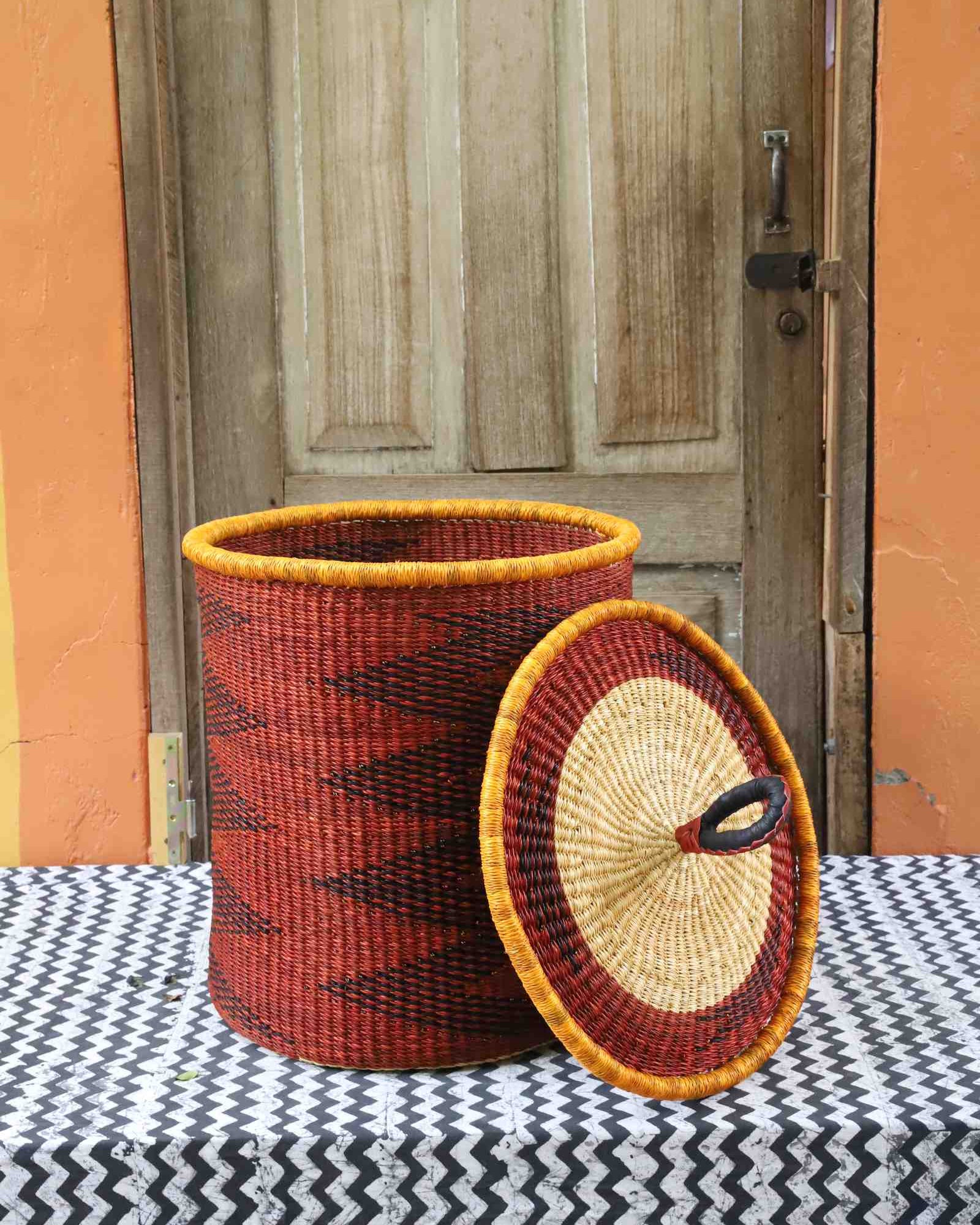 Coiled-rim laundry basket in rust weave with a black diamond pattern, lid resting alongside on a chevron cloth.