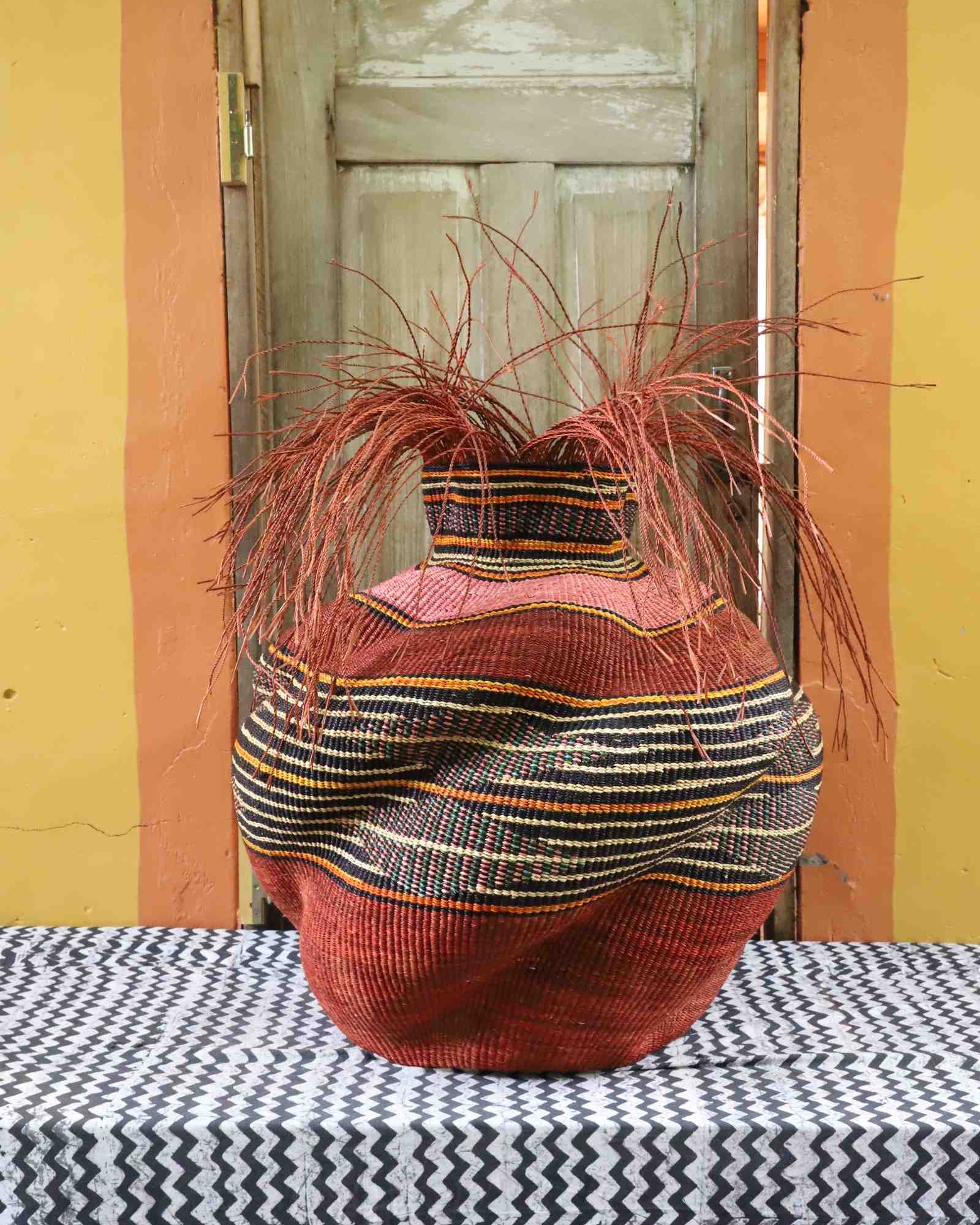 Large round Bolga basket in rust red with a black, cream and orange striped band and loose straw fronds at the neck.