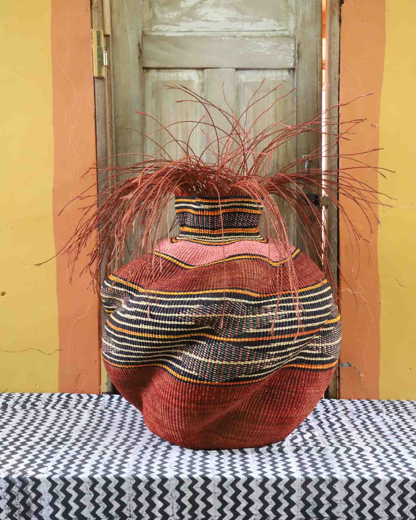 Round Bolga basket in rust red with a black and orange striped band, its narrow neck topped with loose straw fronds.