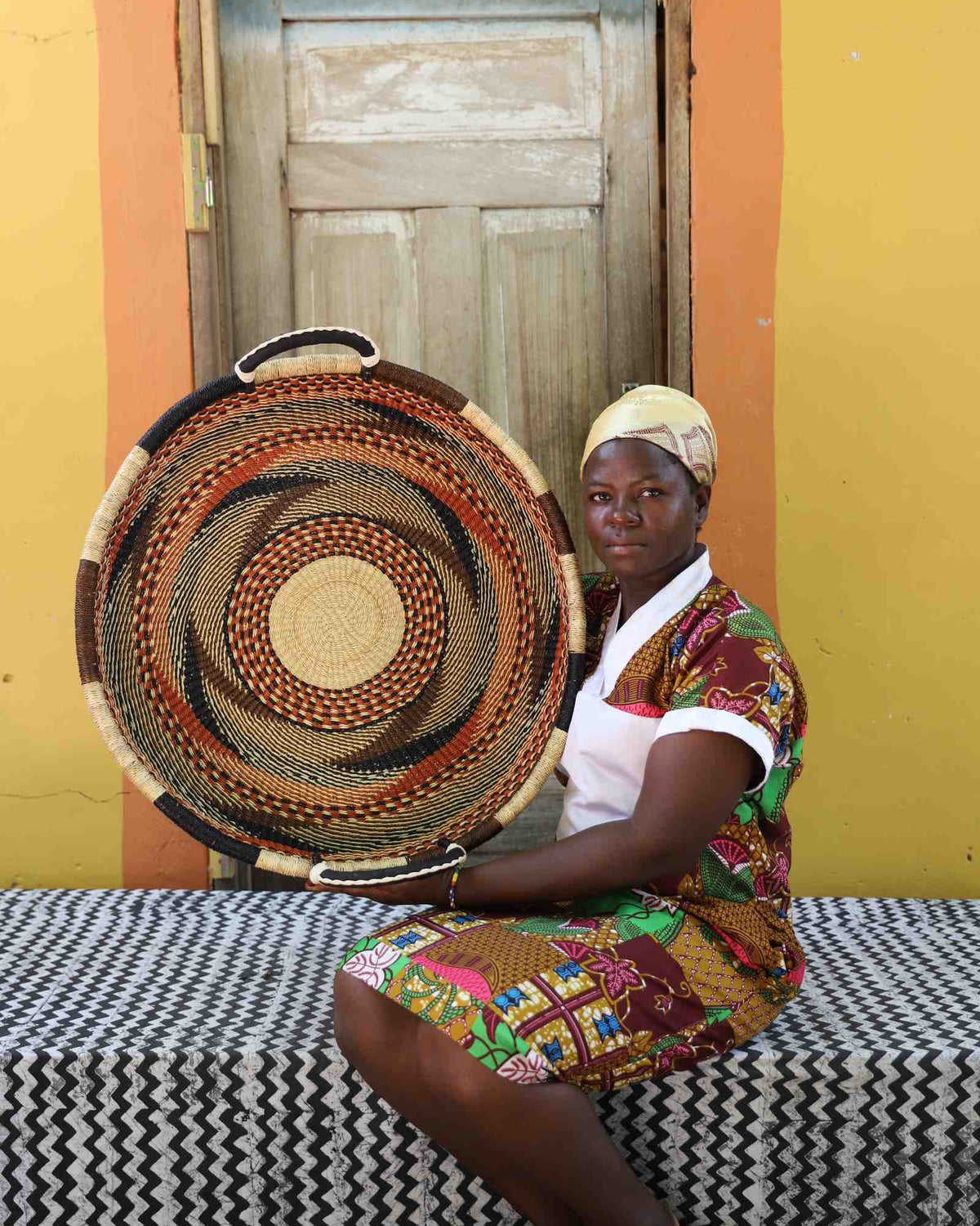 Medium handwoven Bolga wall tray disc with brown and orange bands, held by a seated woman in a printed dress.