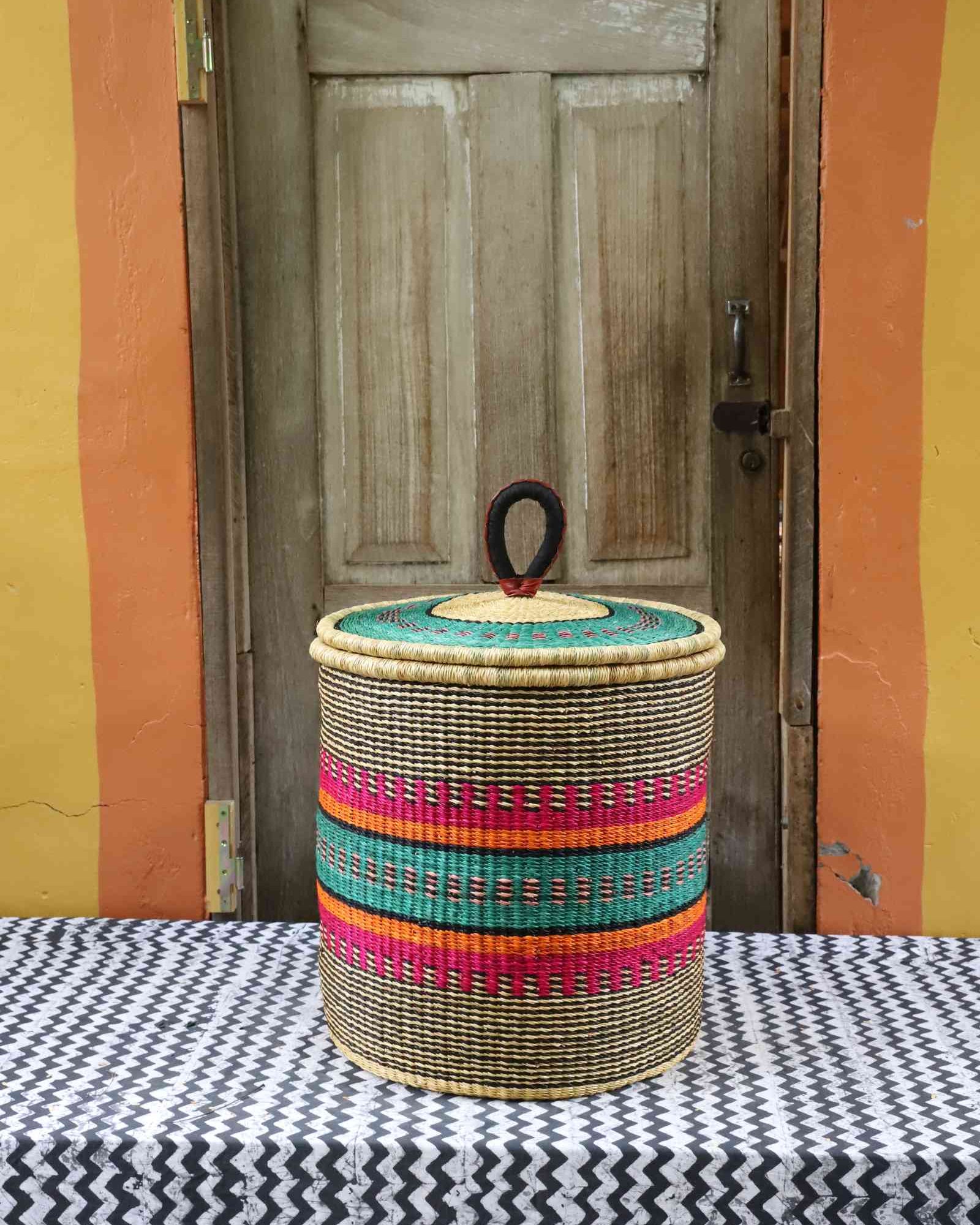 Handwoven Bolga laundry basket with coiled rim and lid in natural straw with pink, orange, green and teal fine stripes.