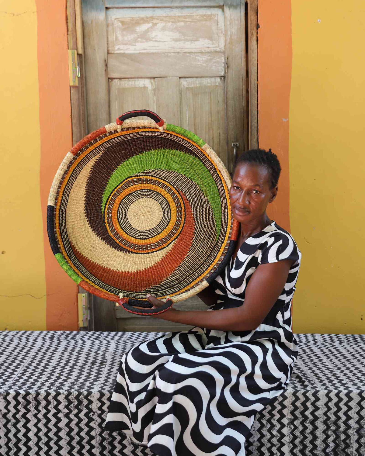 Medium handwoven Bolga wall tray disc with green and orange swirl, held by a seated woman in stripes.
