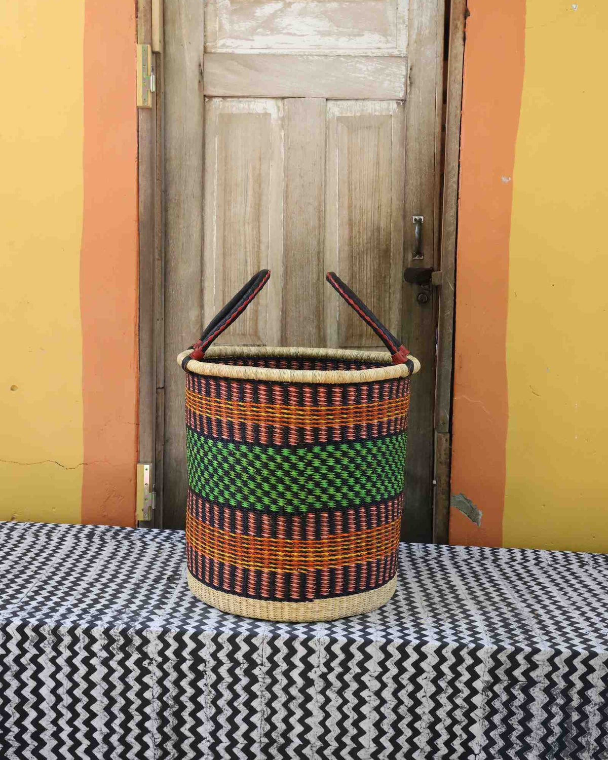 Large handwoven open-top Bolga laundry basket in dark straw with a green central band and orange stripes.