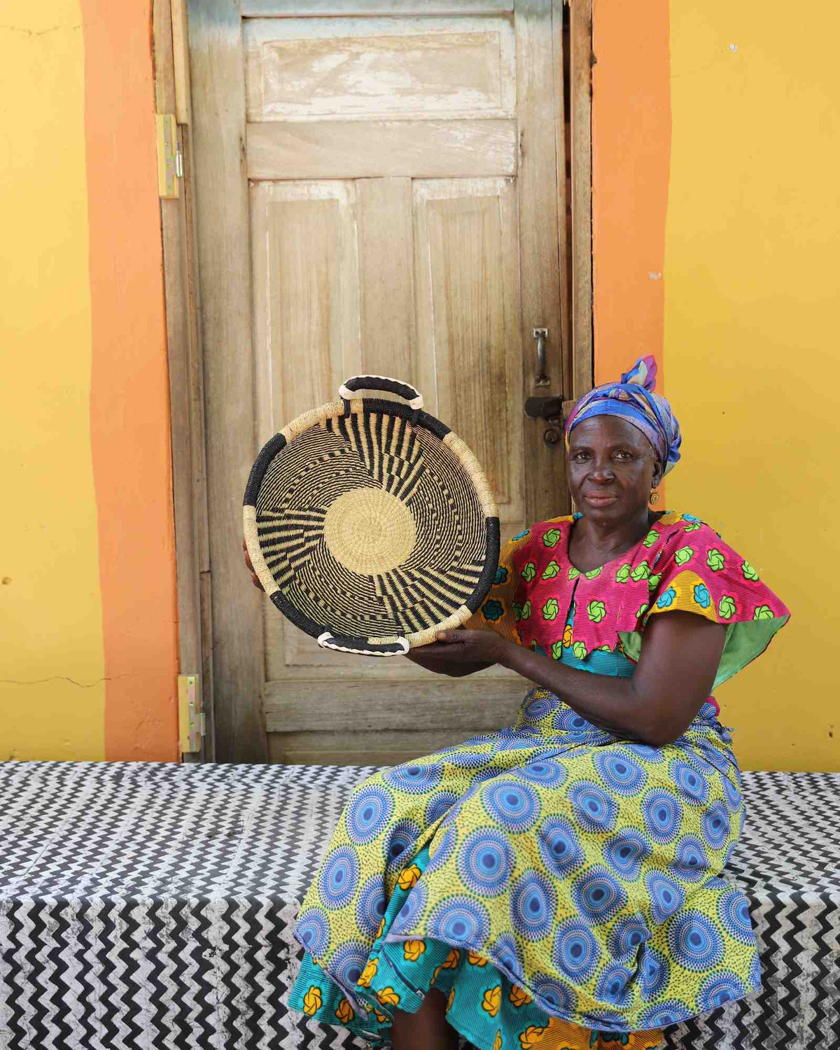 Handwoven Bolga wall tray disc in dark straw with fine radial stripes, held by a seated woman in patterned dress.