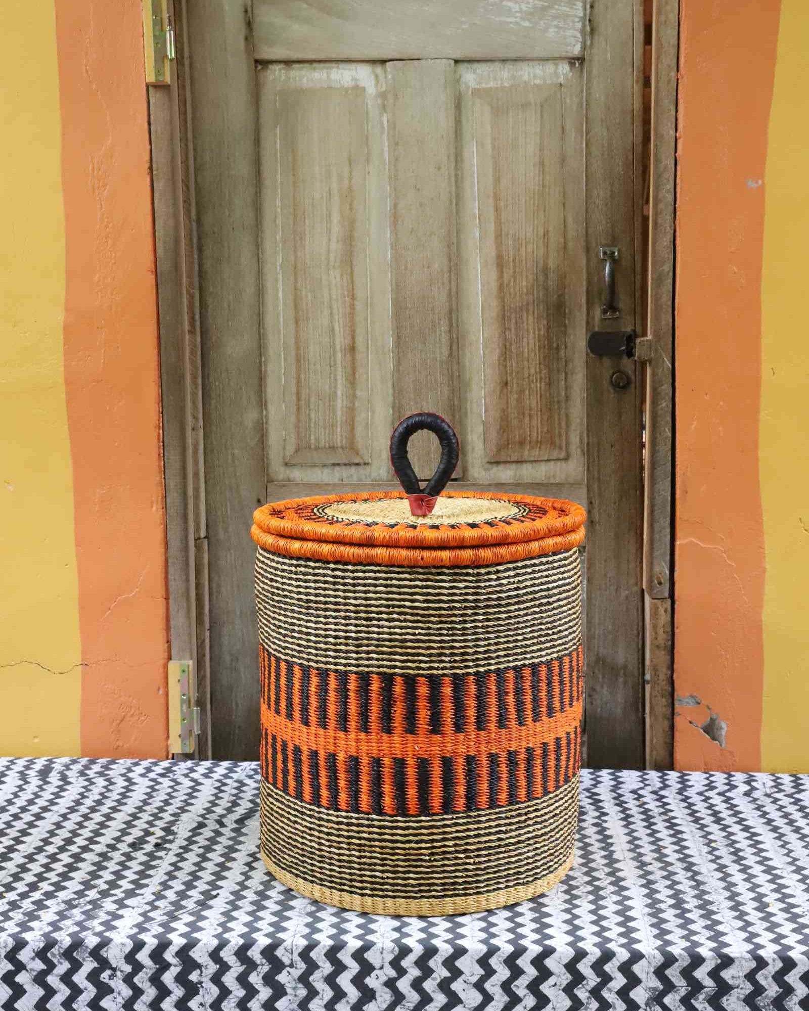 Handwoven Bolga laundry basket with coiled rim and lid in natural straw with a bold orange banded stripe.