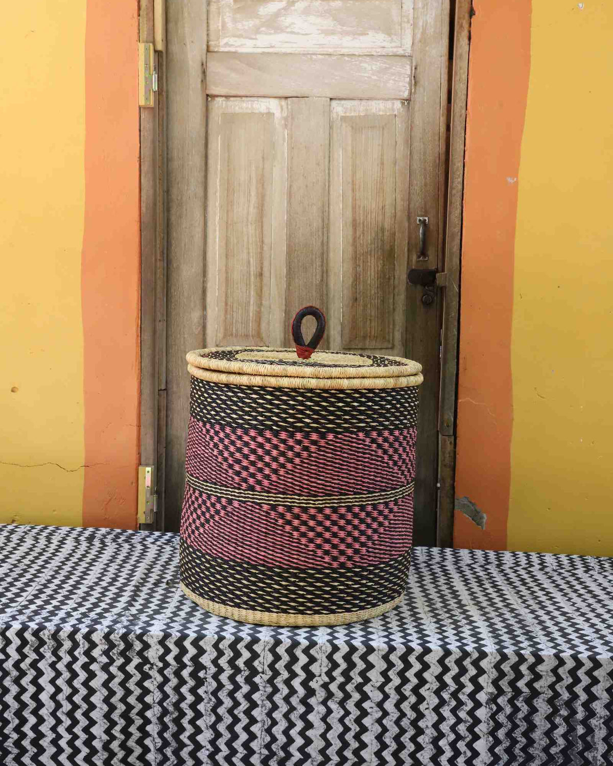 Handwoven Bolga laundry basket with coiled rim and lid in natural straw with red-pink checkered central band.