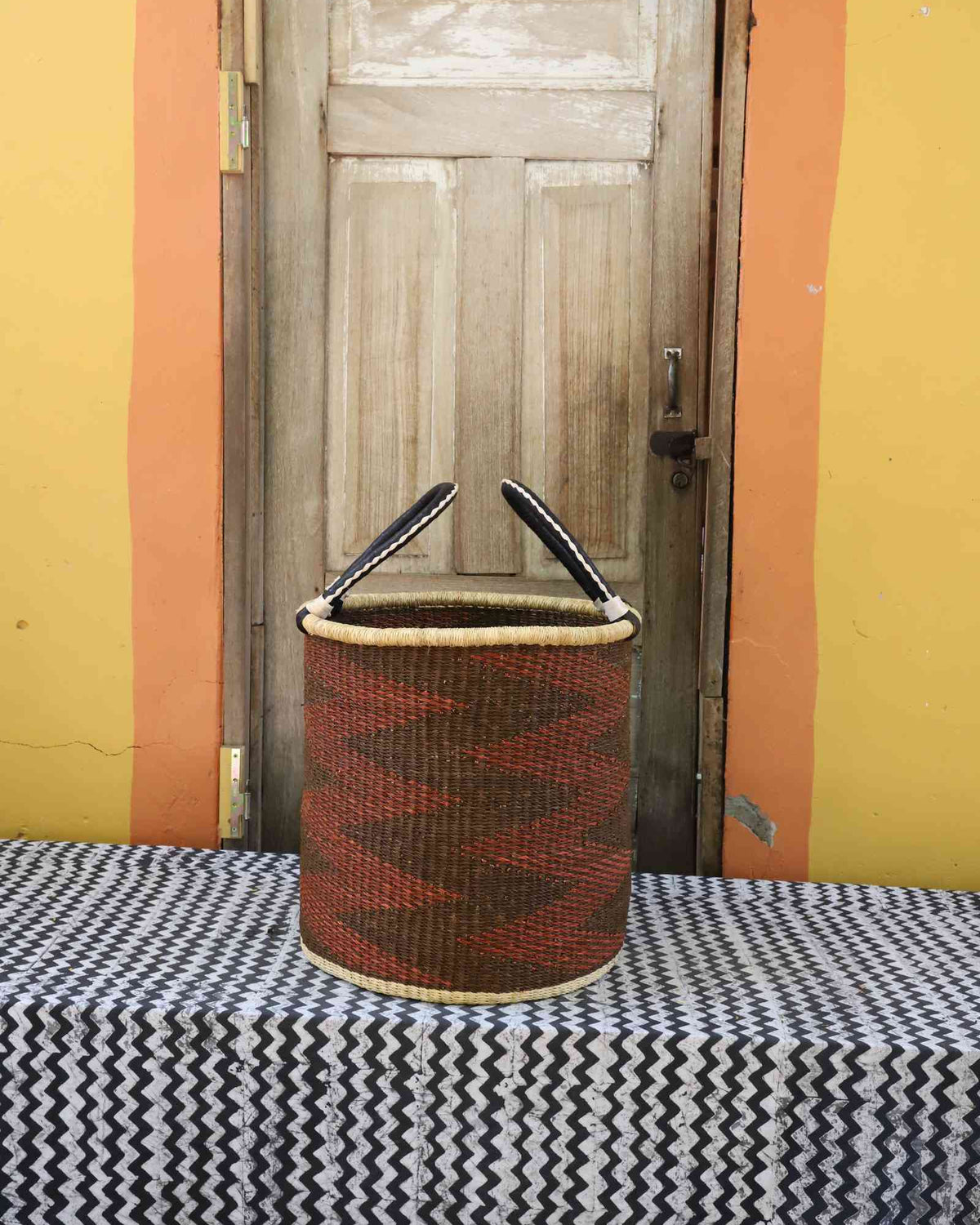 Medium handwoven open-top Bolga laundry basket in rust-orange with dark diagonal zigzag stripes.