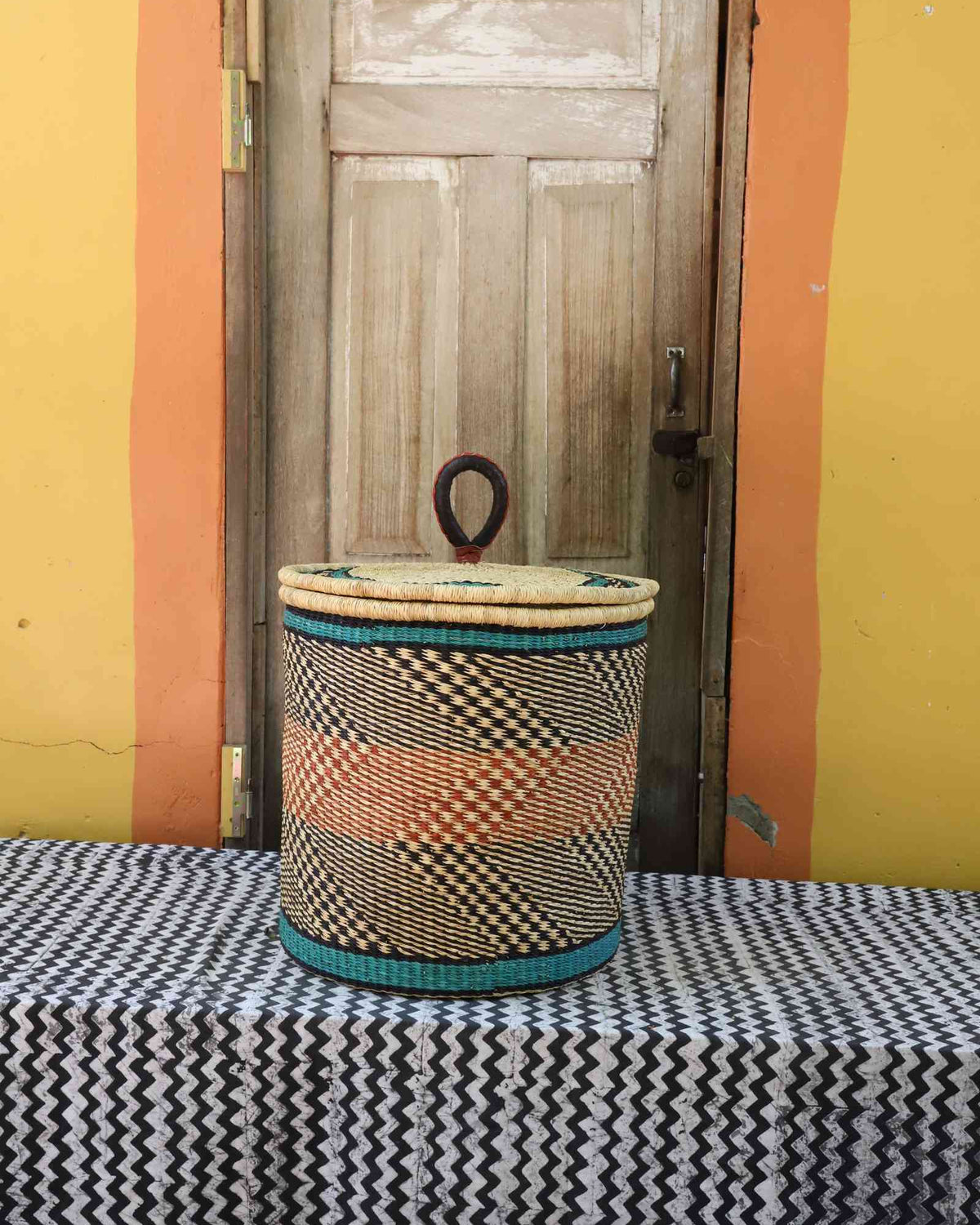 Handwoven Bolga laundry basket with coiled rim and lid in natural straw with orange and dark banded stripes.