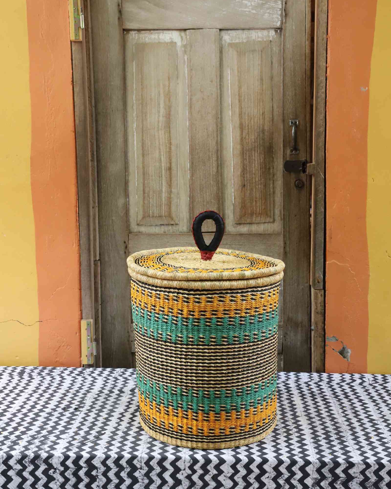 Handwoven Bolga laundry basket with coiled rim and lid in natural straw with orange and teal horizontal bands.