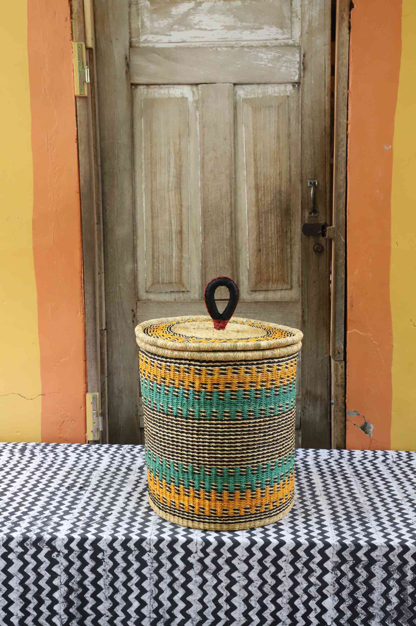 Handwoven Bolga laundry basket with coiled rim and lid in natural straw with orange and teal horizontal bands.