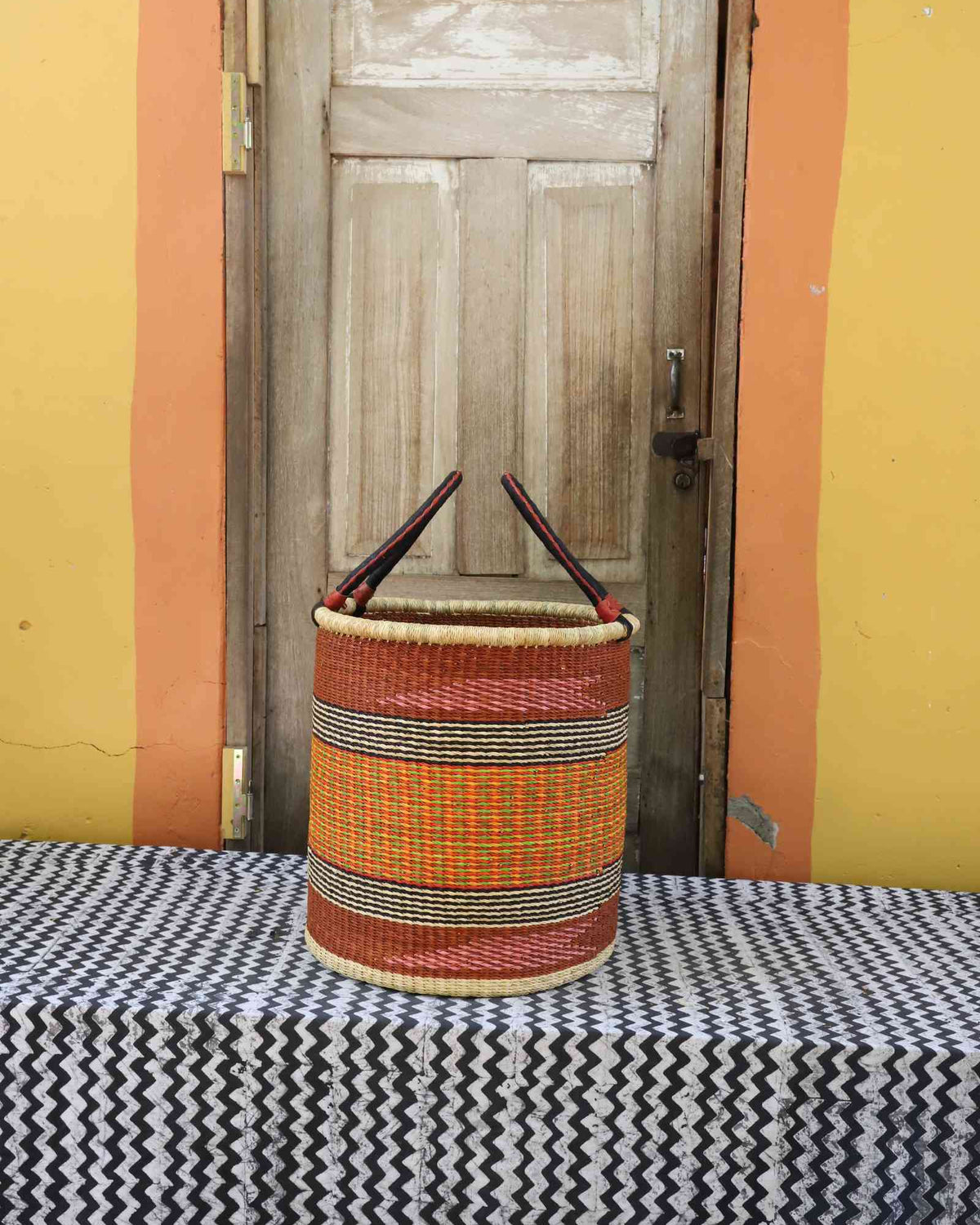 Medium handwoven open-top Bolga laundry basket in natural straw with orange, yellow and red fine bands.